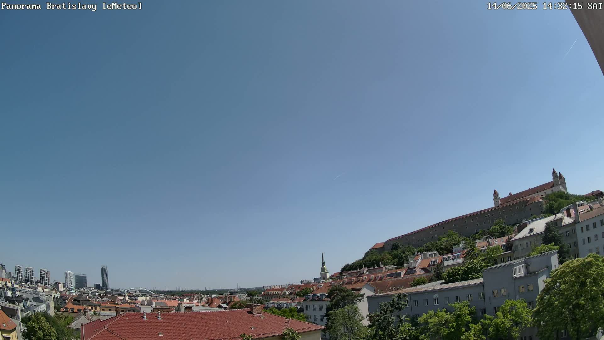 A panoramic view of Bratislava on a clear, sunny day shows a cityscape with red-tiled roofs, green trees, and a large castle on a hill in the background.