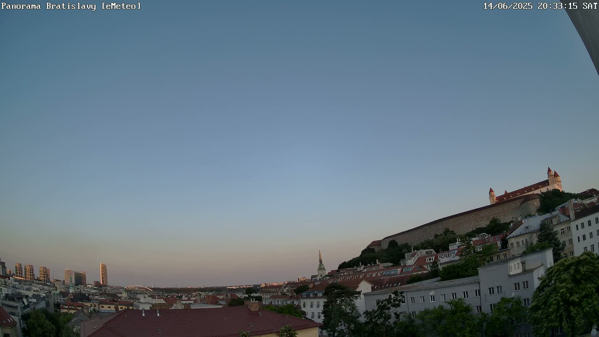 A panoramic view of Bratislava on a clear, sunny day shows a cityscape with red-tiled roofs, green trees, and a large castle on a hill in the background.