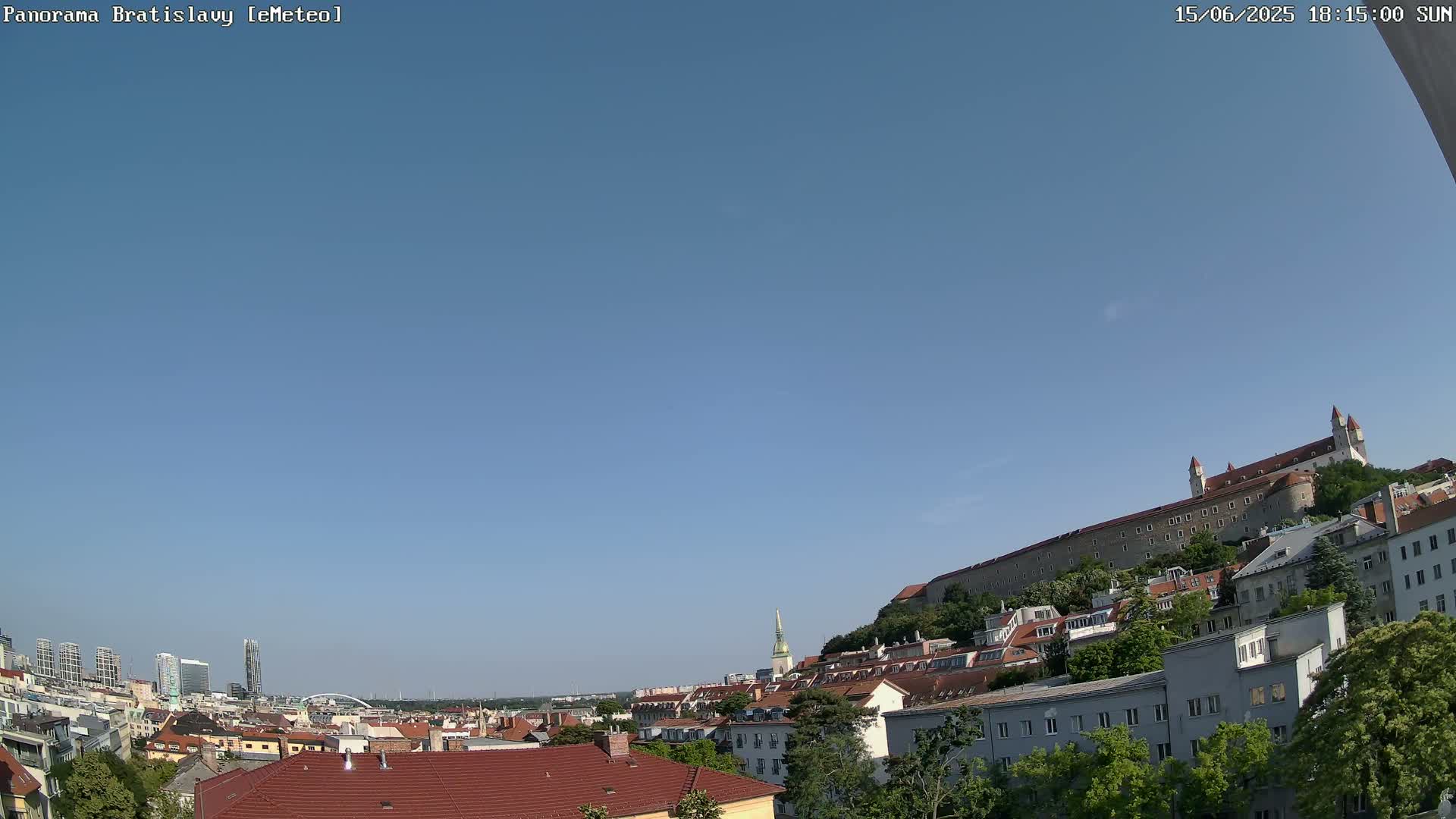 A panoramic view of Bratislava under a clear blue sky shows a cityscape with red-tiled roofs, green trees, and a large stone castle on a hill in the background.