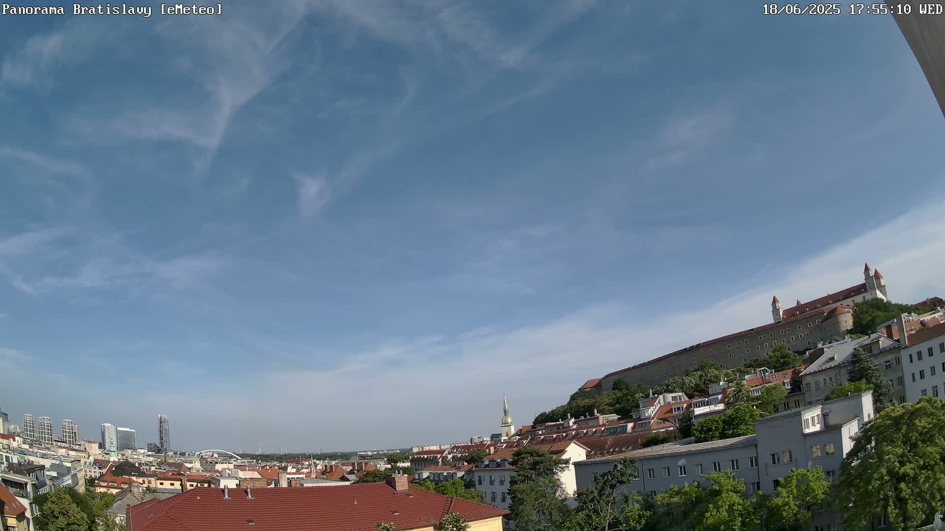 A panoramic view of Bratislava under a mostly clear, bright blue sky shows a cityscape with red-tiled roofs and a large castle atop a hill.