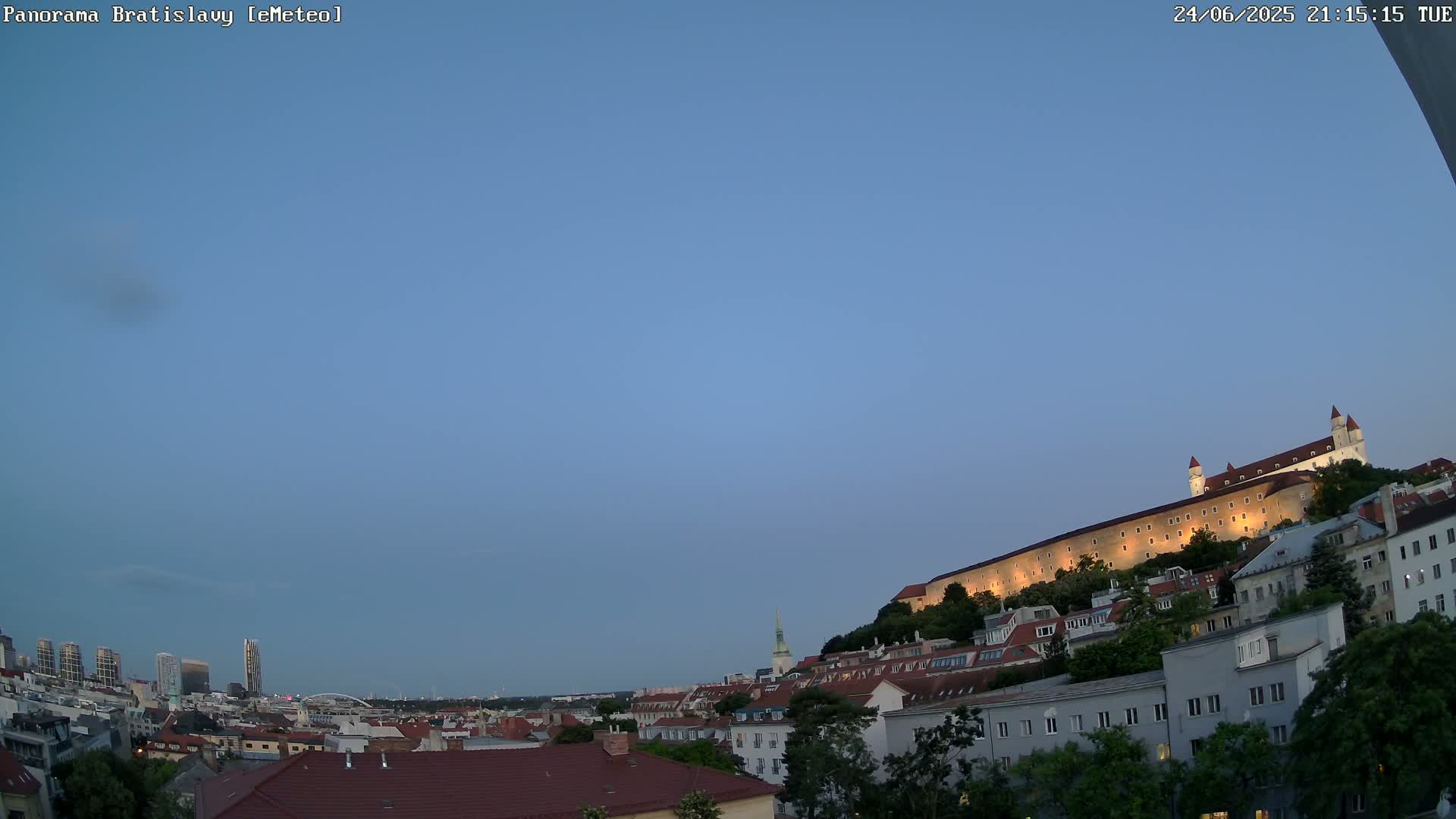 A city skyline at dusk, featuring a large castle illuminated on a hill overlooking a collection of buildings under a clear blue sky.