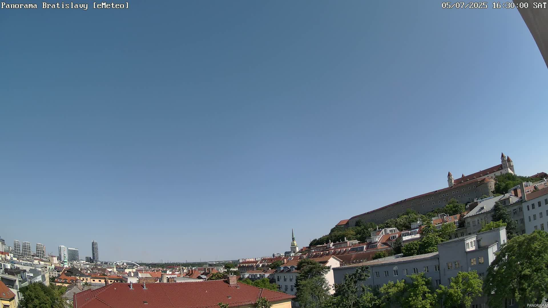 A mostly clear blue sky overlooks a cityscape with red-tiled roofs, interspersed with green trees, culminating in a large, grey stone castle on a hill.