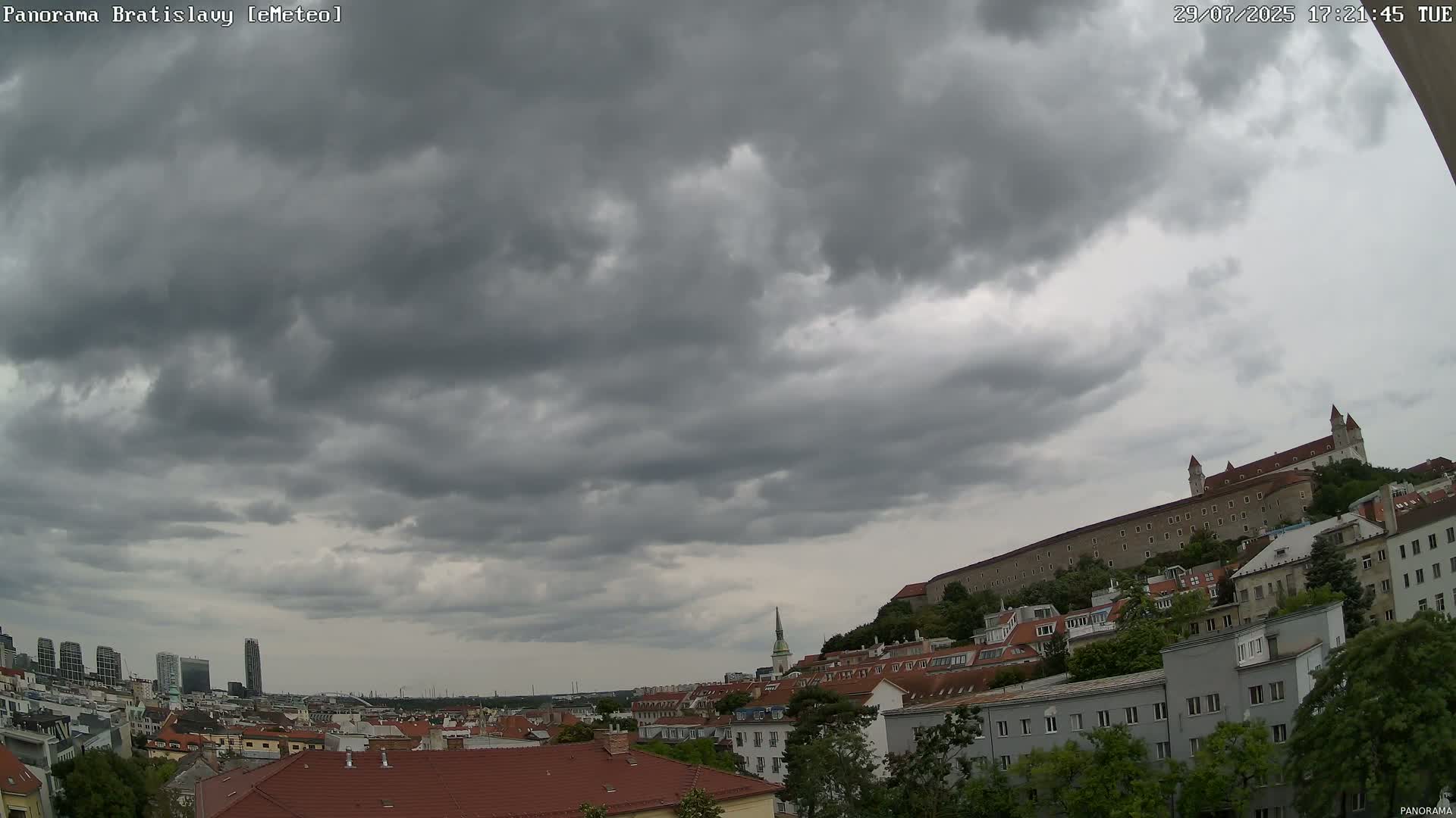 A partly cloudy sky overlooks a city with a large castle on a hill, featuring buildings with red tile roofs.