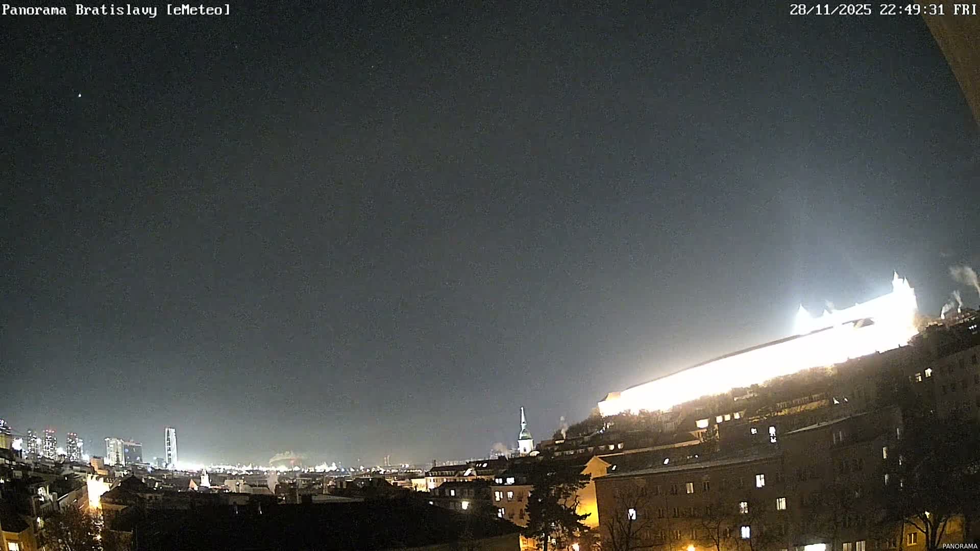 A clear, dark night sky hangs over a panoramic cityscape featuring a brightly illuminated castle atop a hill, a church spire, residential buildings with glowing windows, and a distant cluster of modern high-rises.