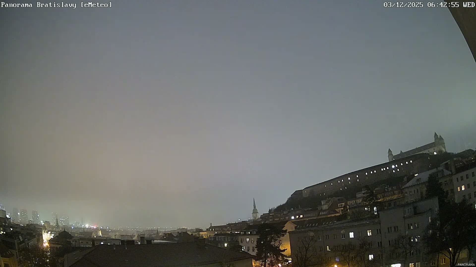 A sprawling city panorama at dawn is enveloped in dense fog, with an illuminated castle prominent on a hill to the right, and numerous buildings with lit windows extending towards a hazy distant skyline.