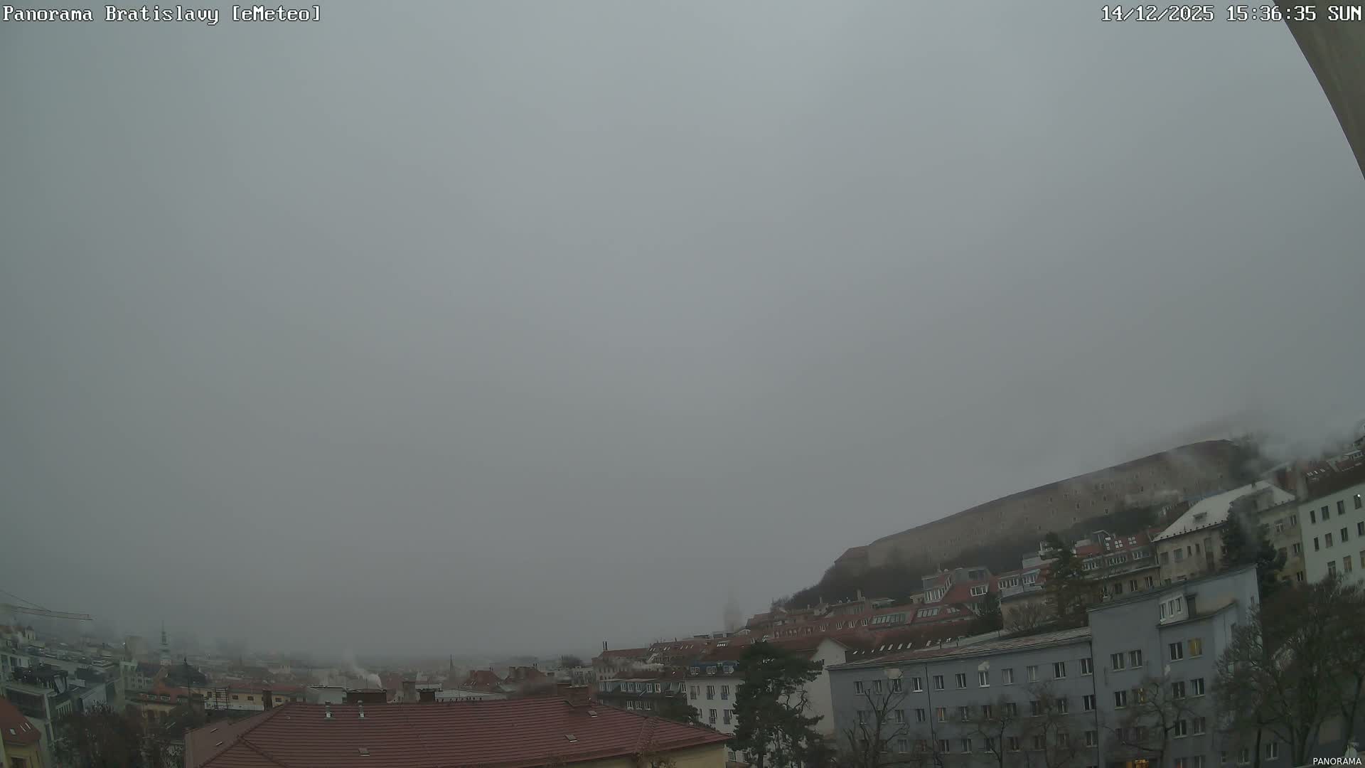 A sprawling city panorama at dawn is enveloped in dense fog, with an illuminated castle prominent on a hill to the right, and numerous buildings with lit windows extending towards a hazy distant skyline.