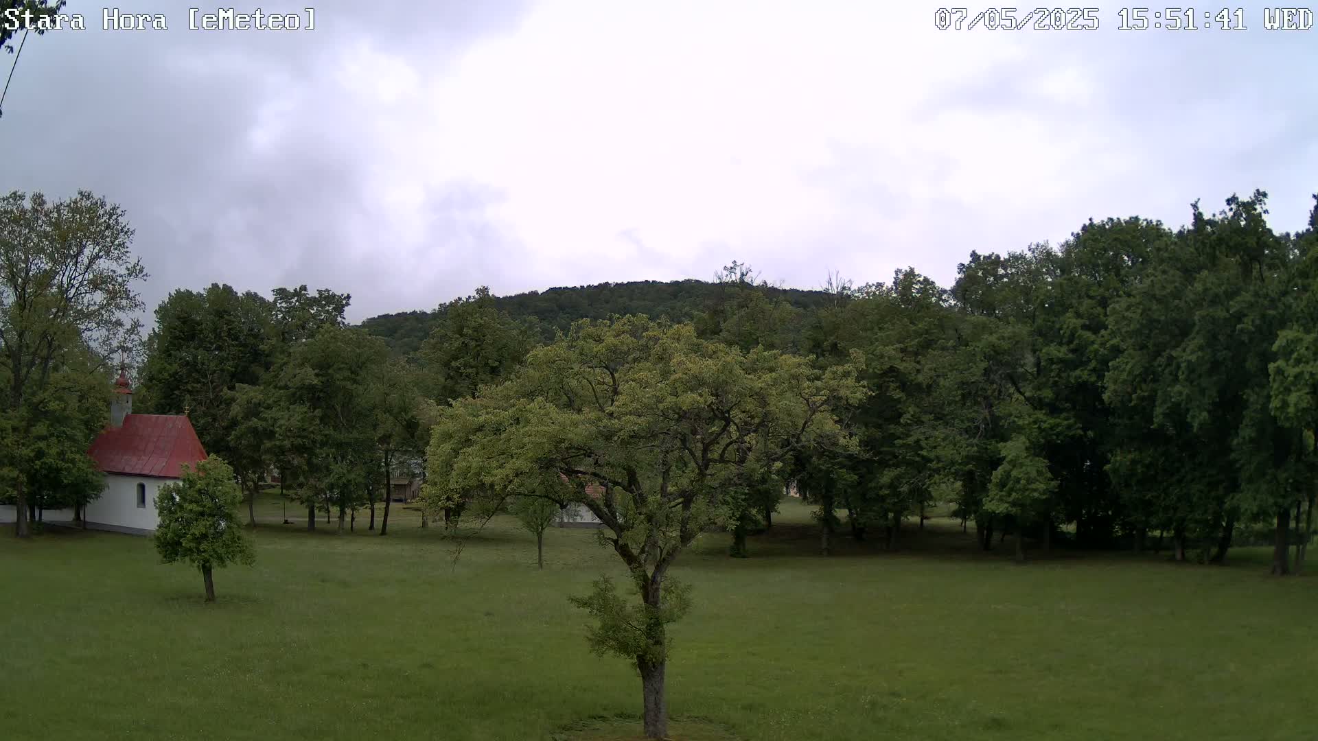 A small white chapel with a red roof sits in a grassy field surrounded by lush green trees, under an overcast sky.