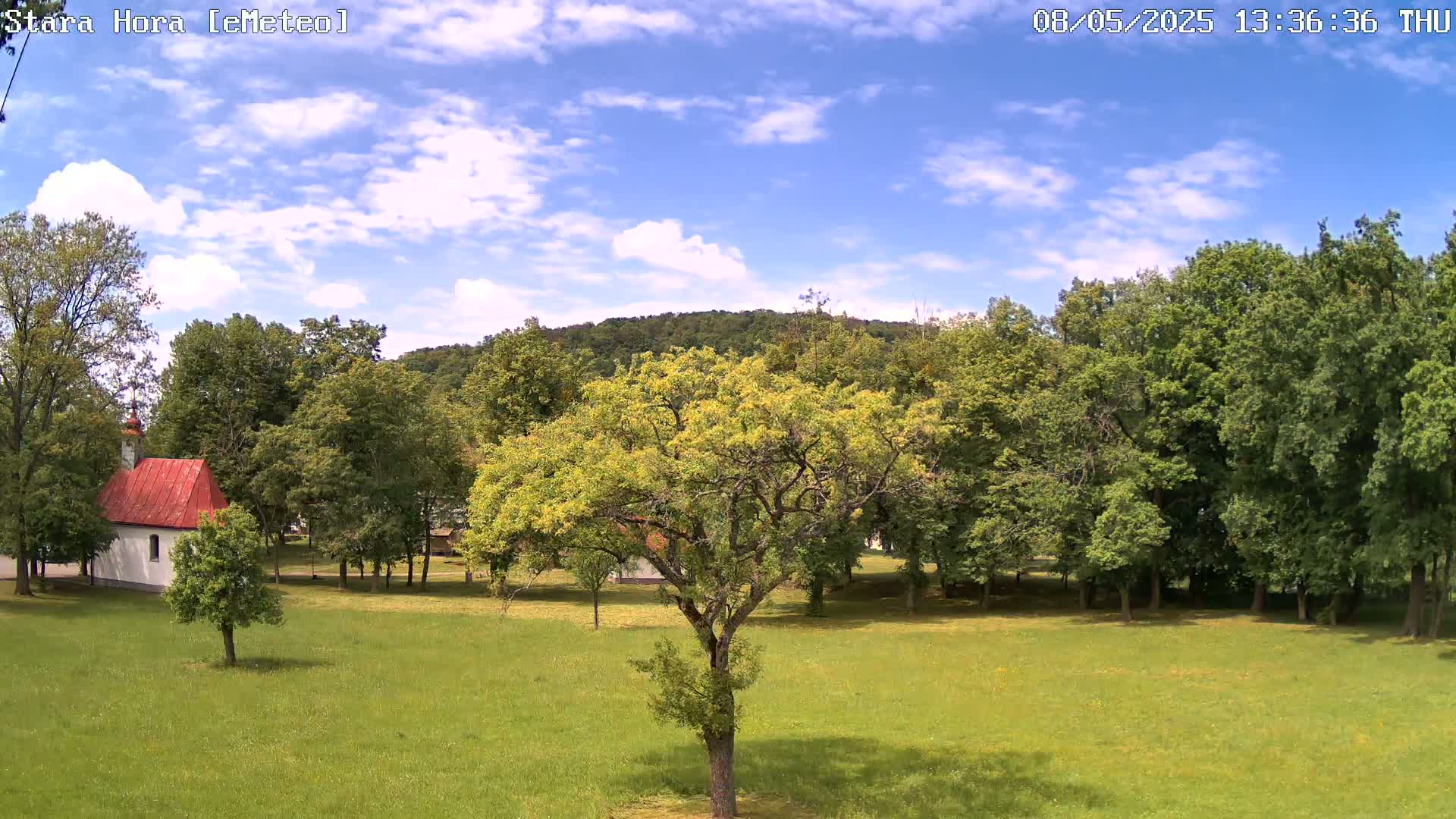 A partly cloudy sky overlooks a grassy field with a small white chapel, several trees, and a forest in the background.