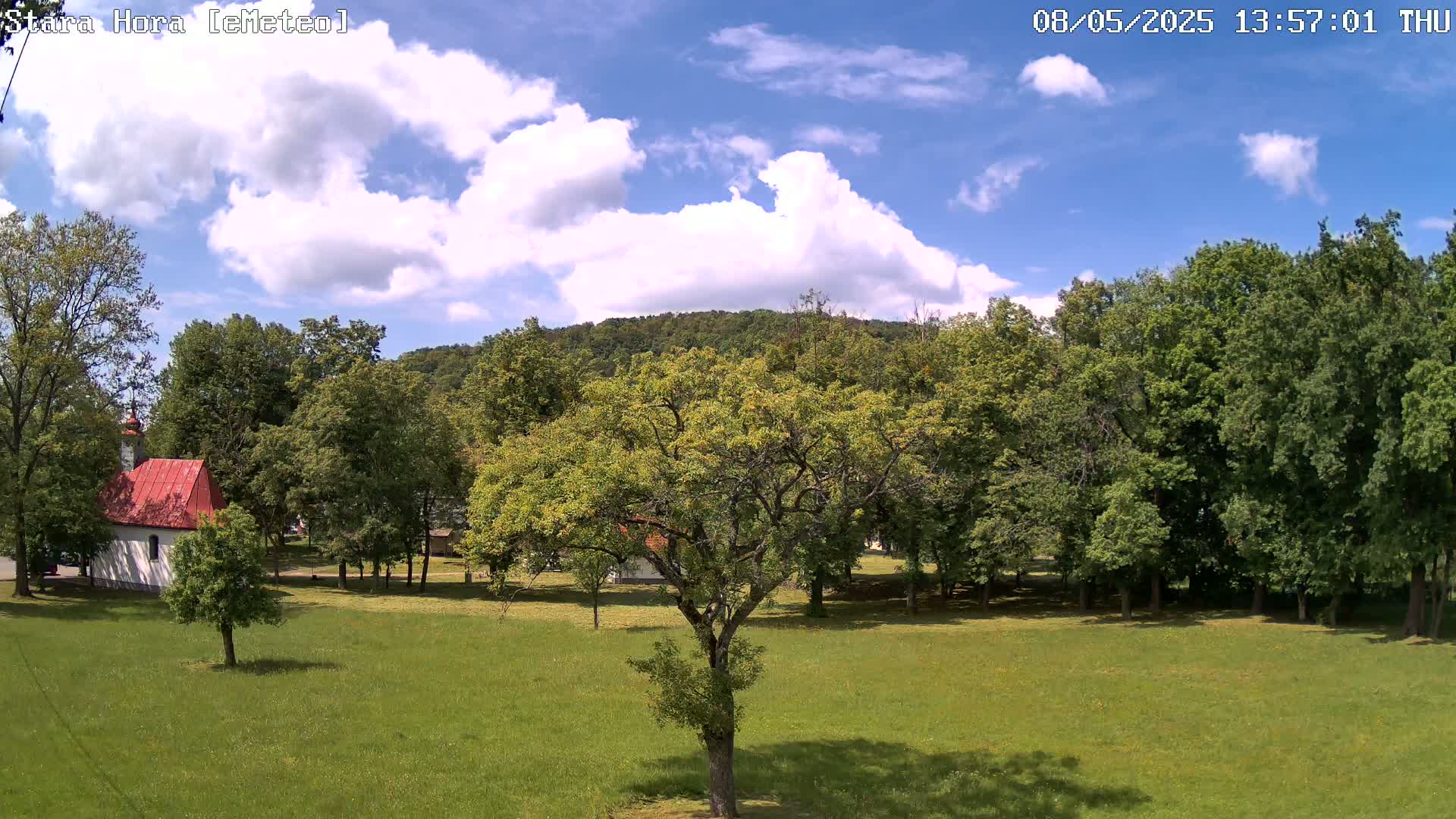 A grassy field with a small red-roofed building and numerous leafy green trees under a partly cloudy blue sky.