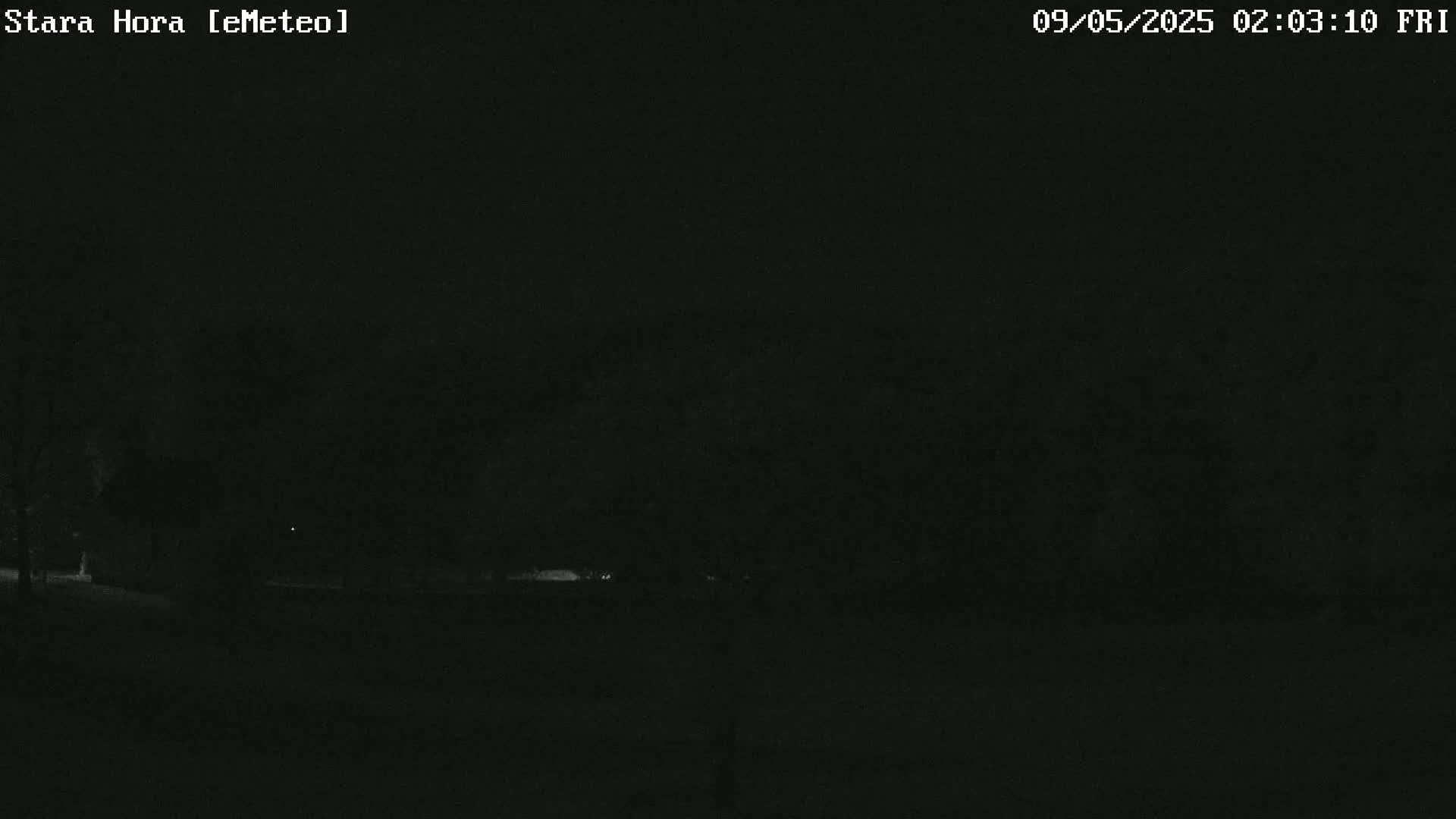 A dark, nighttime scene shows a mostly obscured landscape with some dimly lit trees and a flat, dark foreground.