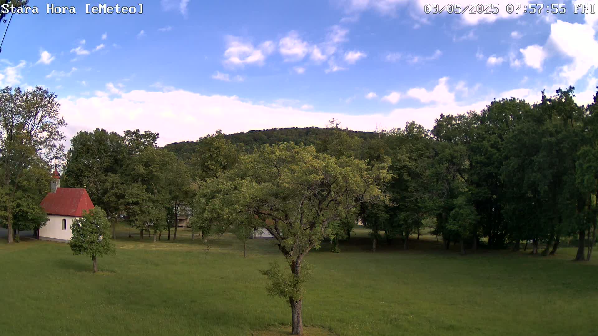 A small white chapel with a red roof sits in a grassy field surrounded by green trees under a partly cloudy blue sky.