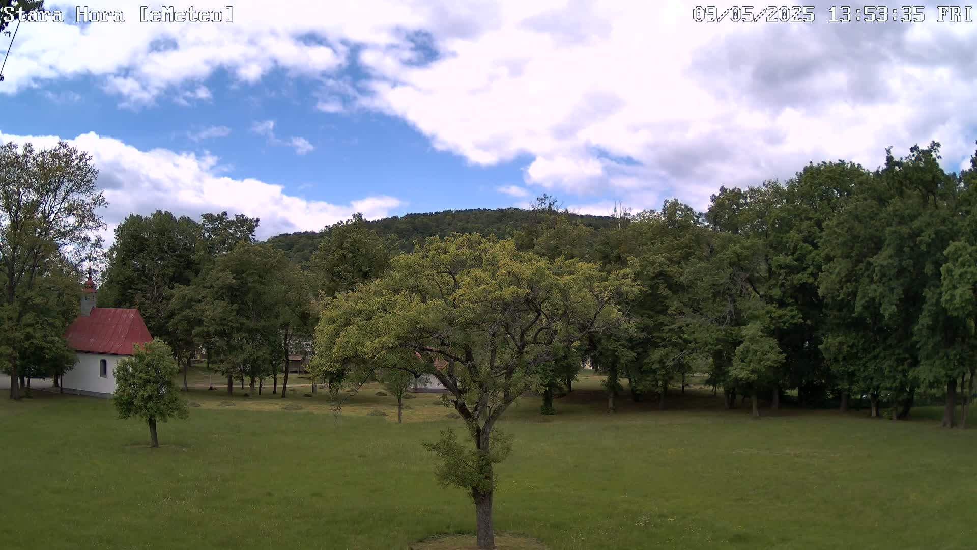 A partly cloudy sky overlooks a green field with a small red-roofed building, several trees, and a hill covered in lush forest in the background.
