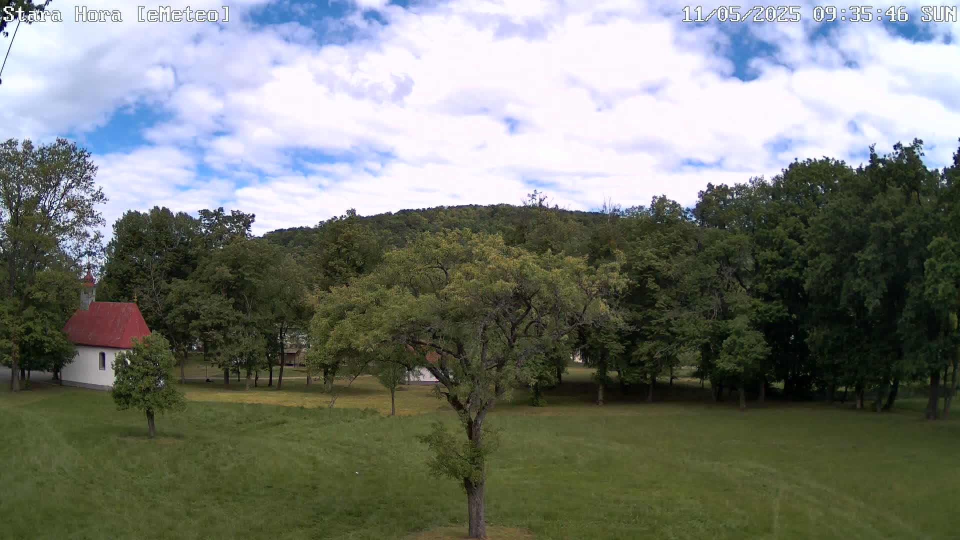 A small white chapel with a red roof sits in a grassy field surrounded by lush green trees and a hill under a partly cloudy sky.