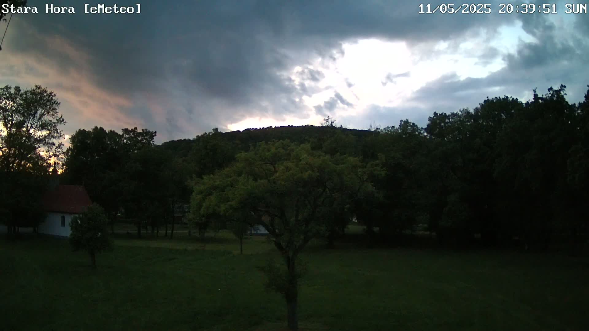 A small white building sits in a grassy field surrounded by dark trees and a hill under a partly cloudy, dusky sky.