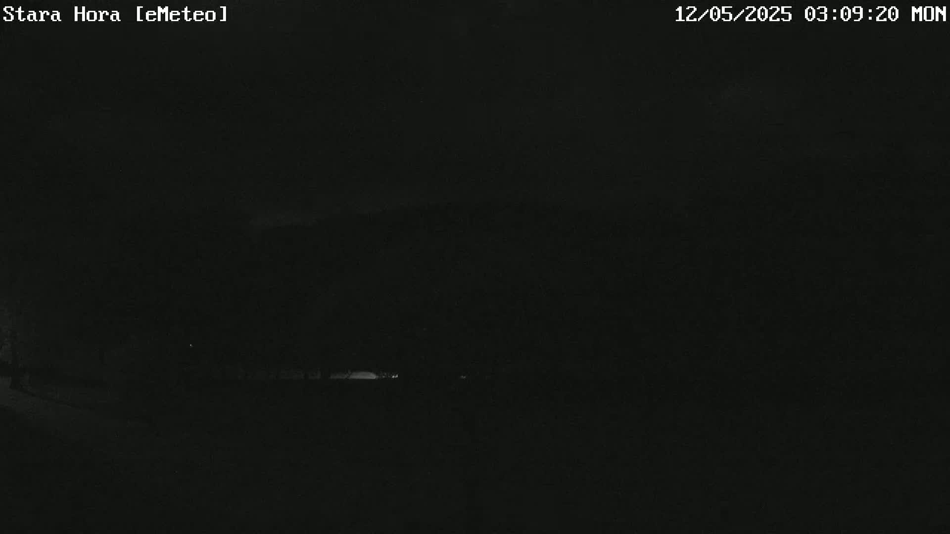 A dark, nighttime outdoor scene shows a dimly lit landscape with a few faint lights in the distance under a mostly clear sky.