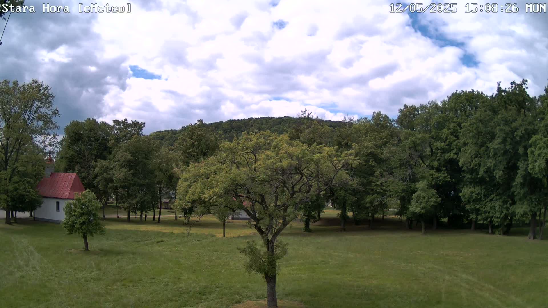 A partly cloudy sky overlooks a grassy field with a small, white building and many green trees, some of which surround a hill in the distance.