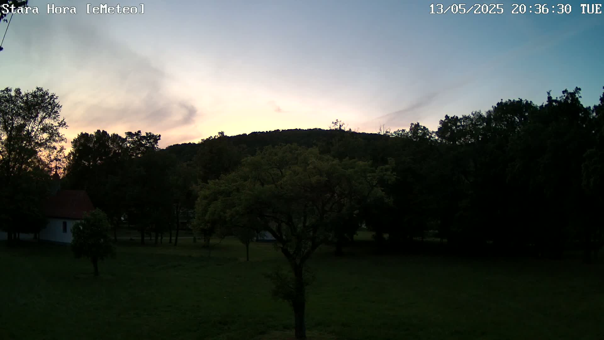 A twilight view shows a small white building, several trees, and a hill under a pale pink and blue sky.