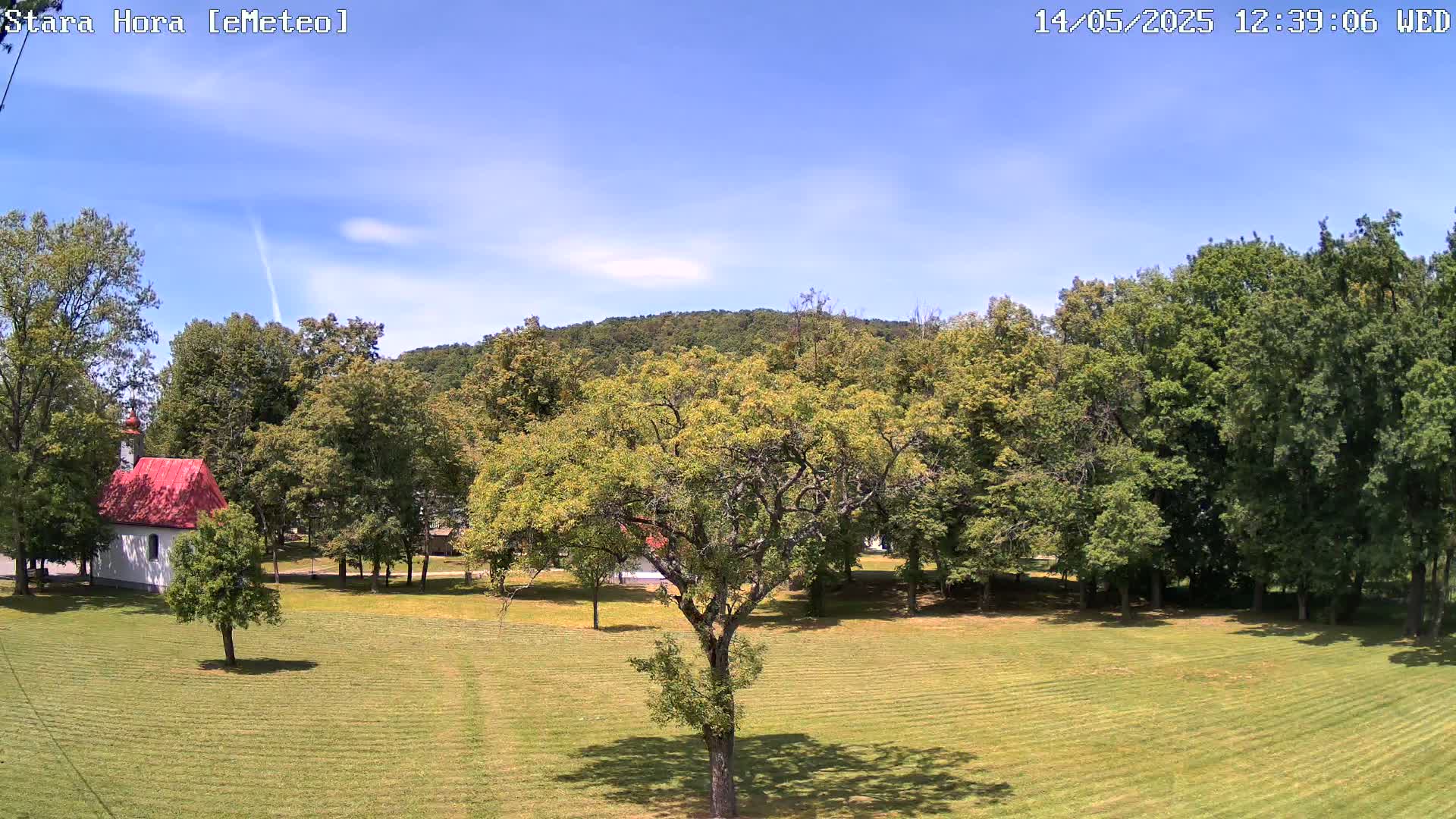 A small red-roofed chapel sits in a grassy field surrounded by lush green trees and a hill under a mostly clear blue sky.