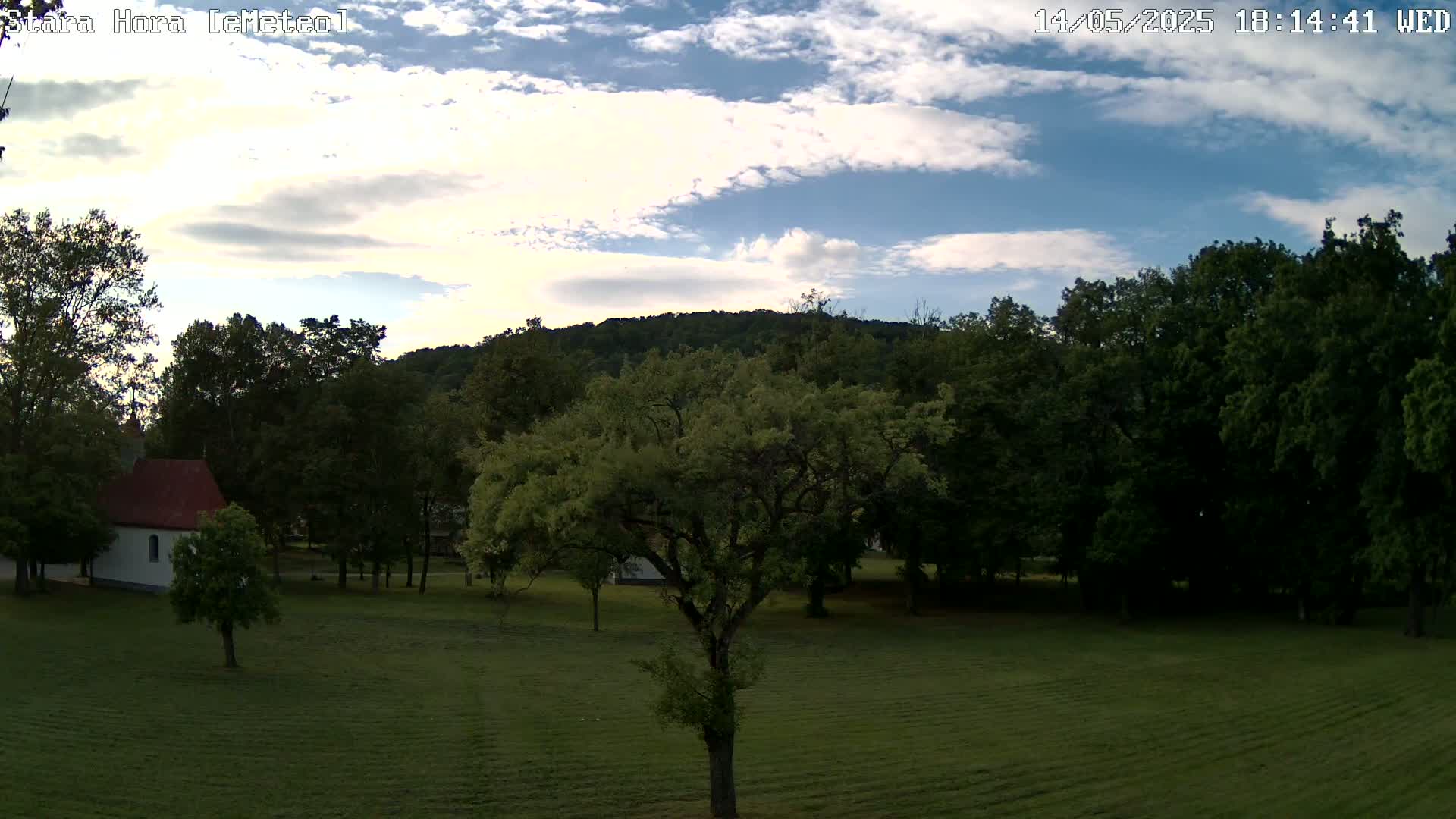 A grassy field with a small white building and many green trees under a partly cloudy sky.