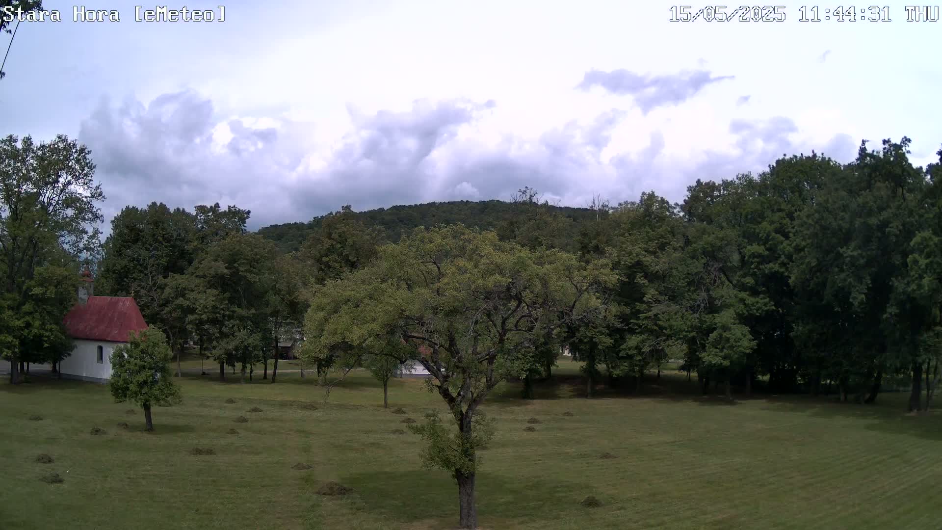 A small white chapel with a red roof sits on a grassy field surrounded by lush green trees and a hill under a partly cloudy sky.