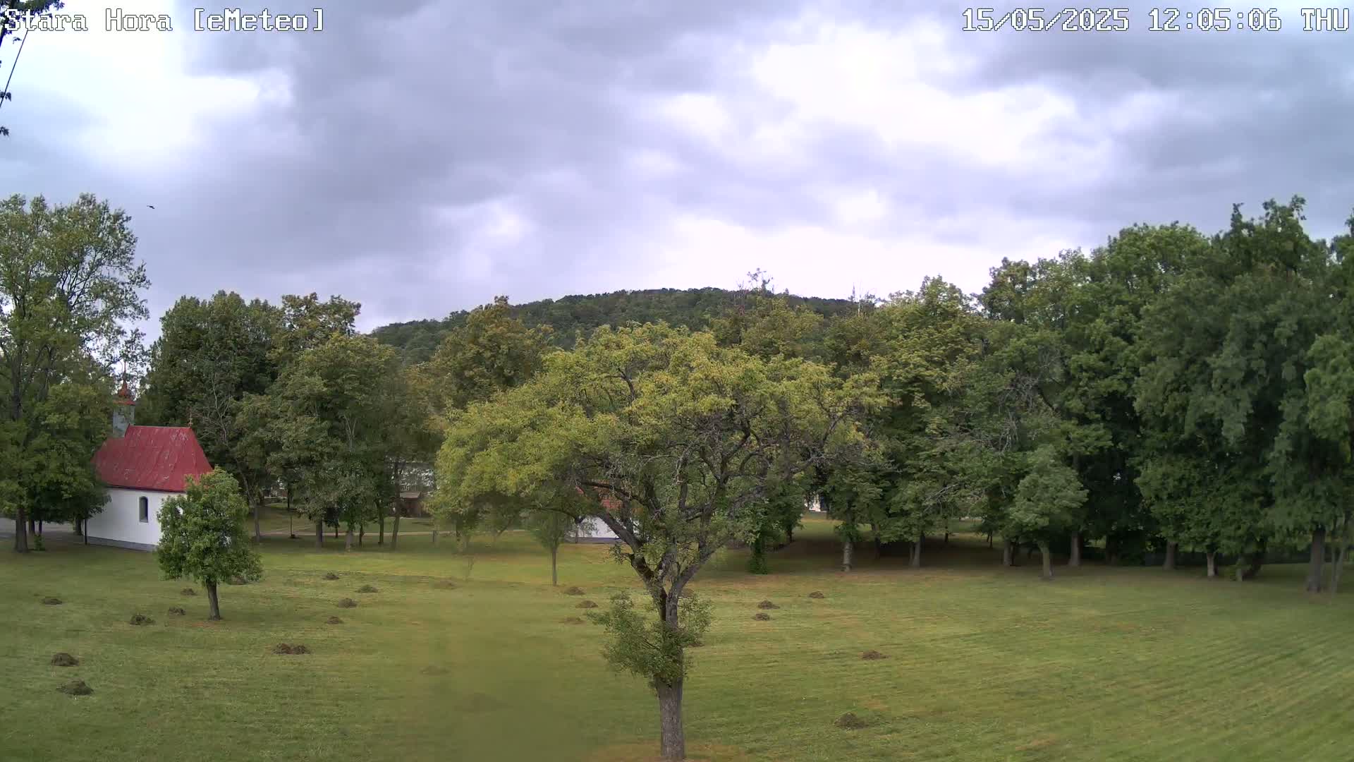 A grassy field with a small white chapel and several leafy green trees under a cloudy sky.