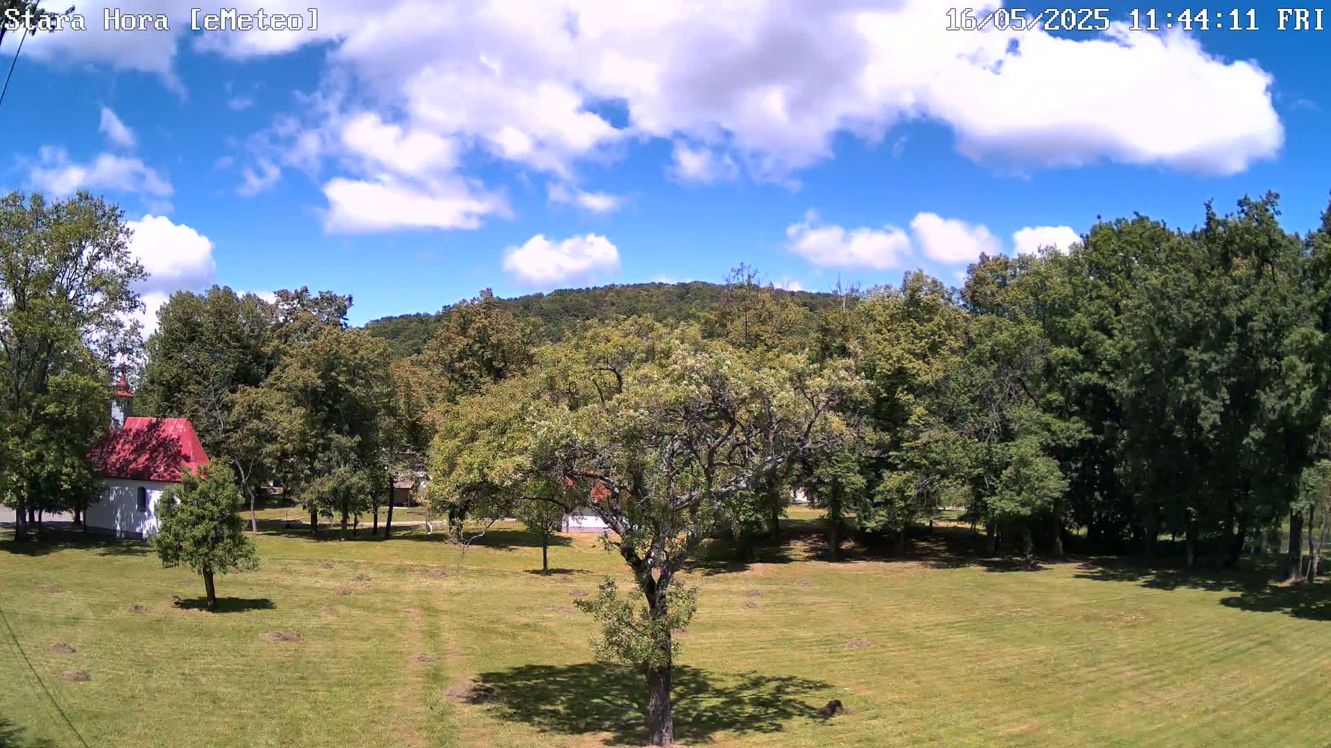 A partly sunny day shows a small white chapel with a red roof in a grassy field surrounded by lush green trees and a hill in the background.