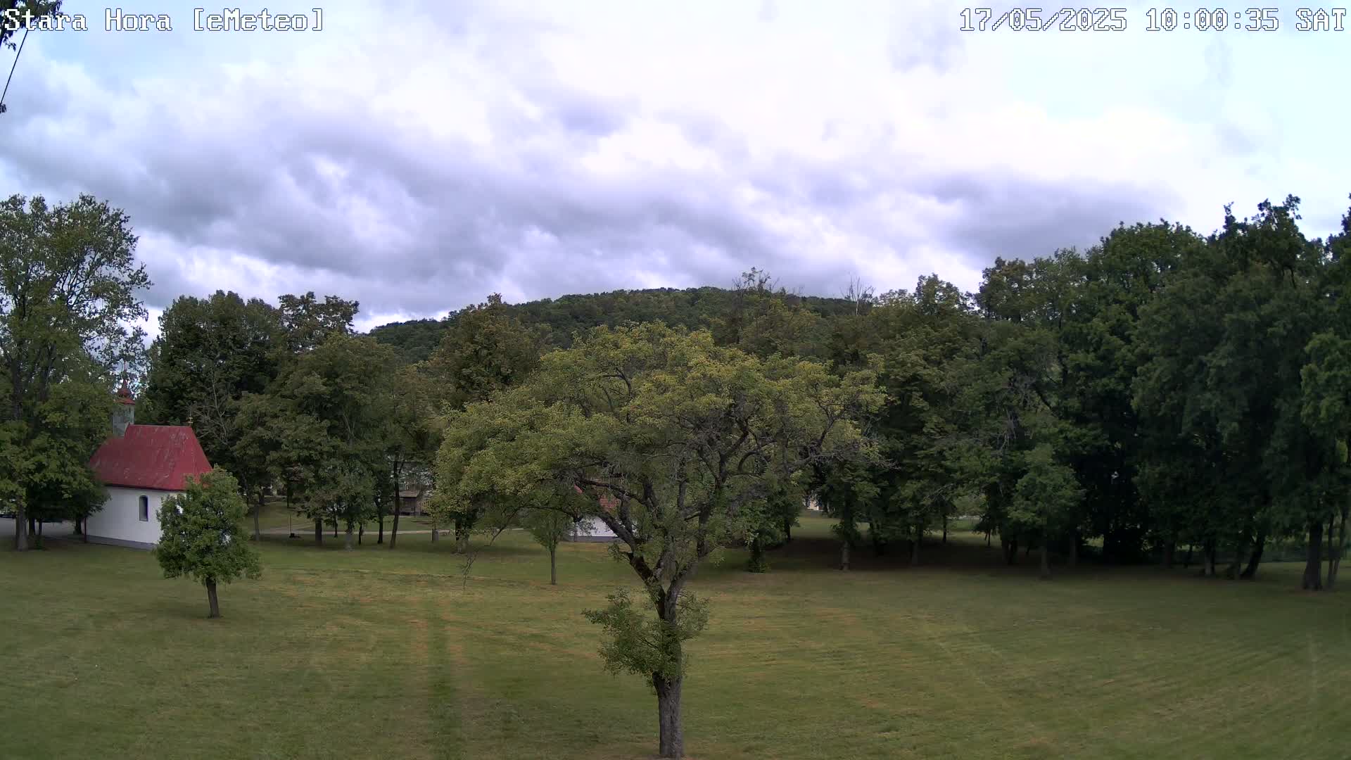 A small white chapel with a red roof sits in a grassy field surrounded by deciduous trees and a hill under a cloudy sky.