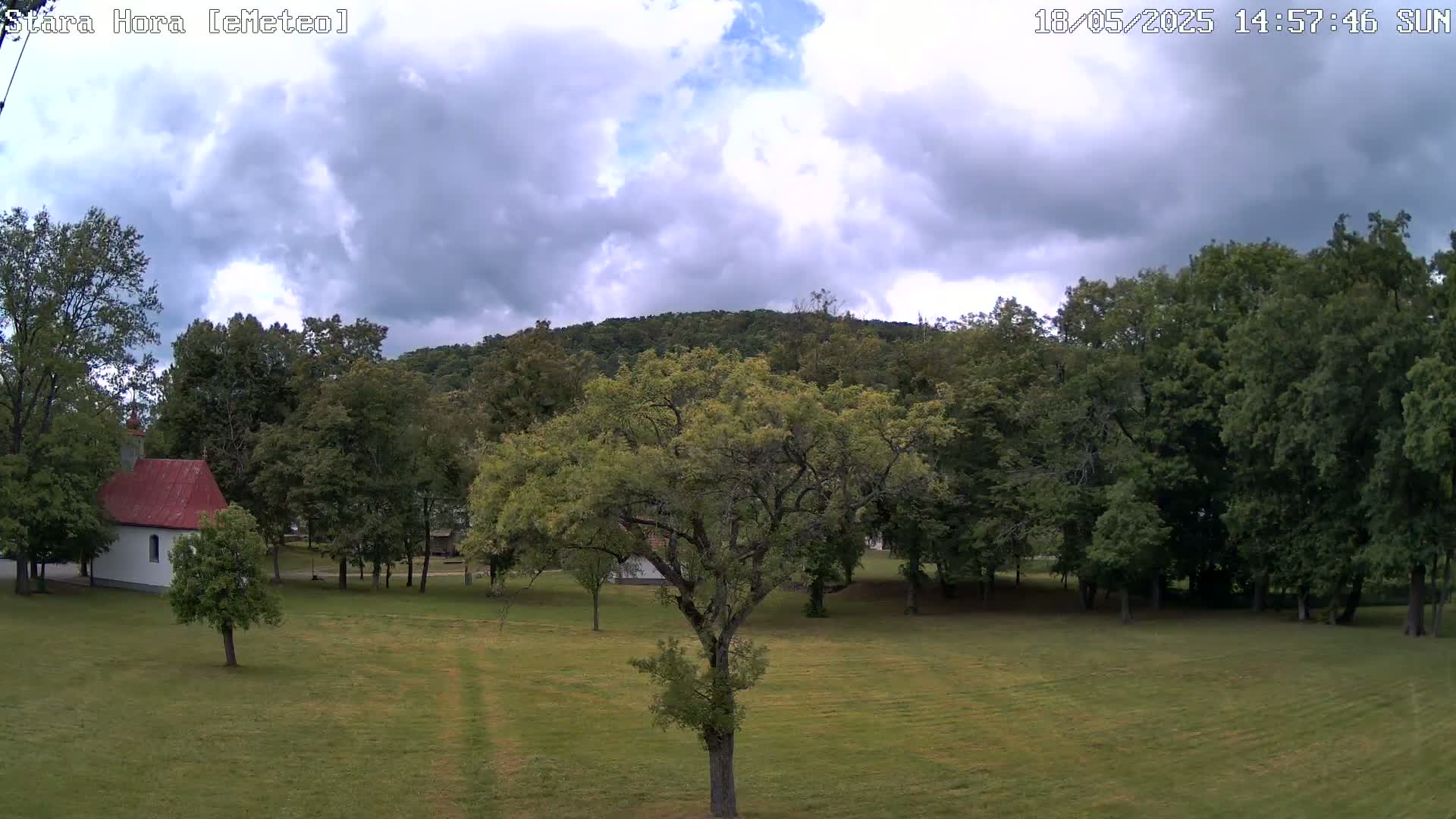 A partly cloudy sky hangs over a grassy field with trees, a small white building with a red roof, and a hill in the background.
