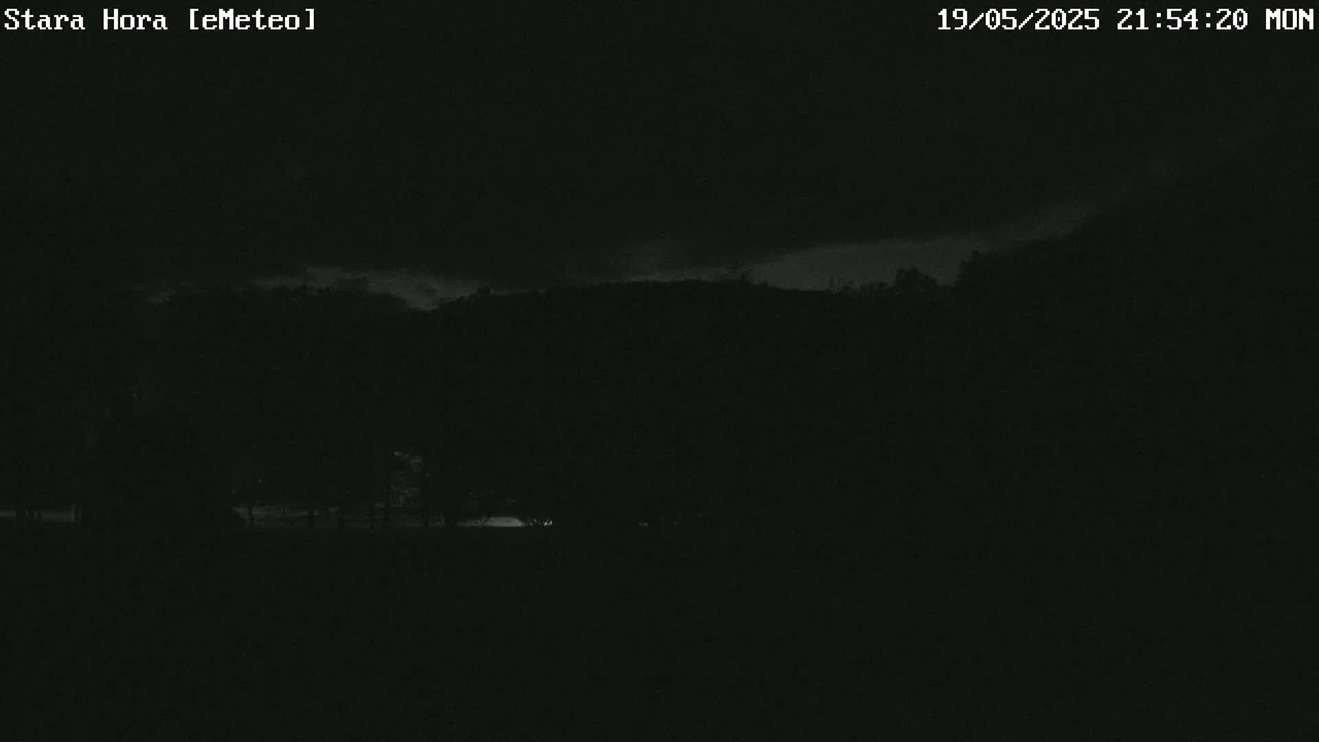 A nighttime, low-light image shows a dark landscape of trees and hills under a mostly cloudy sky.