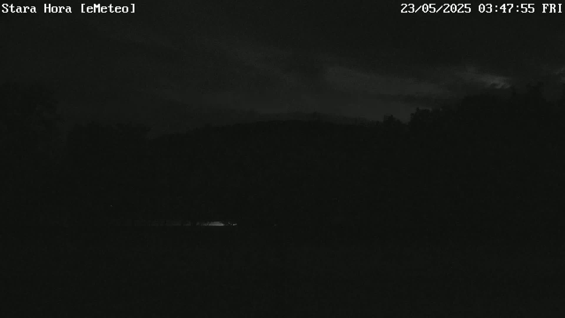 A nighttime, dark image shows a barely visible line of trees and a faint glow near the bottom of the frame under a cloudy sky.