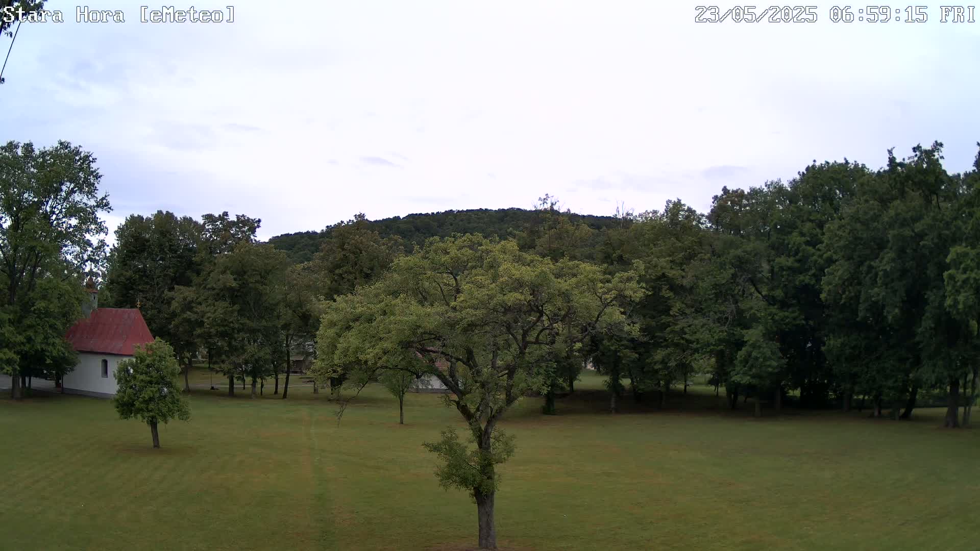 A small white chapel with a red roof sits on a grassy field surrounded by lush green trees and a hill in the background under a mostly cloudy sky.