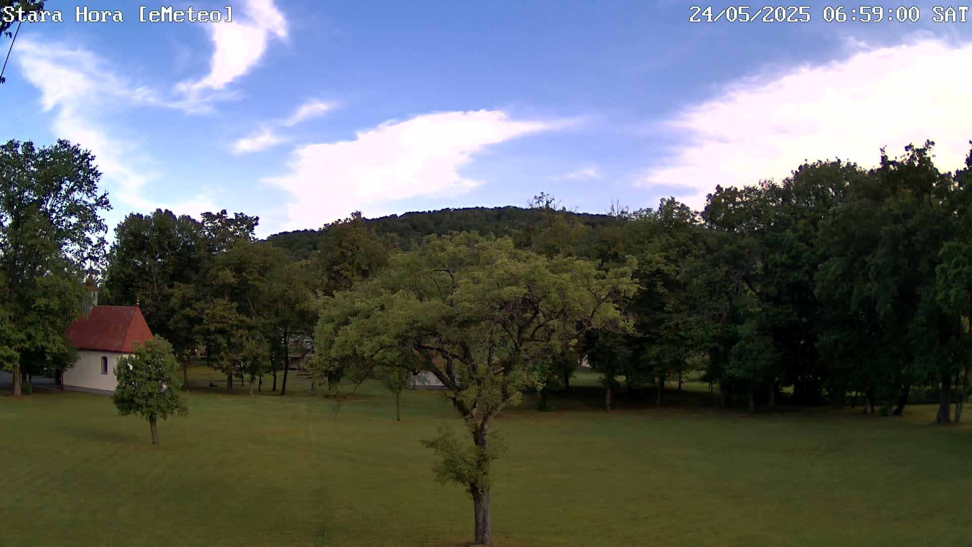 A small white chapel with a red roof sits in a grassy field surrounded by lush green trees under a partly cloudy blue sky.