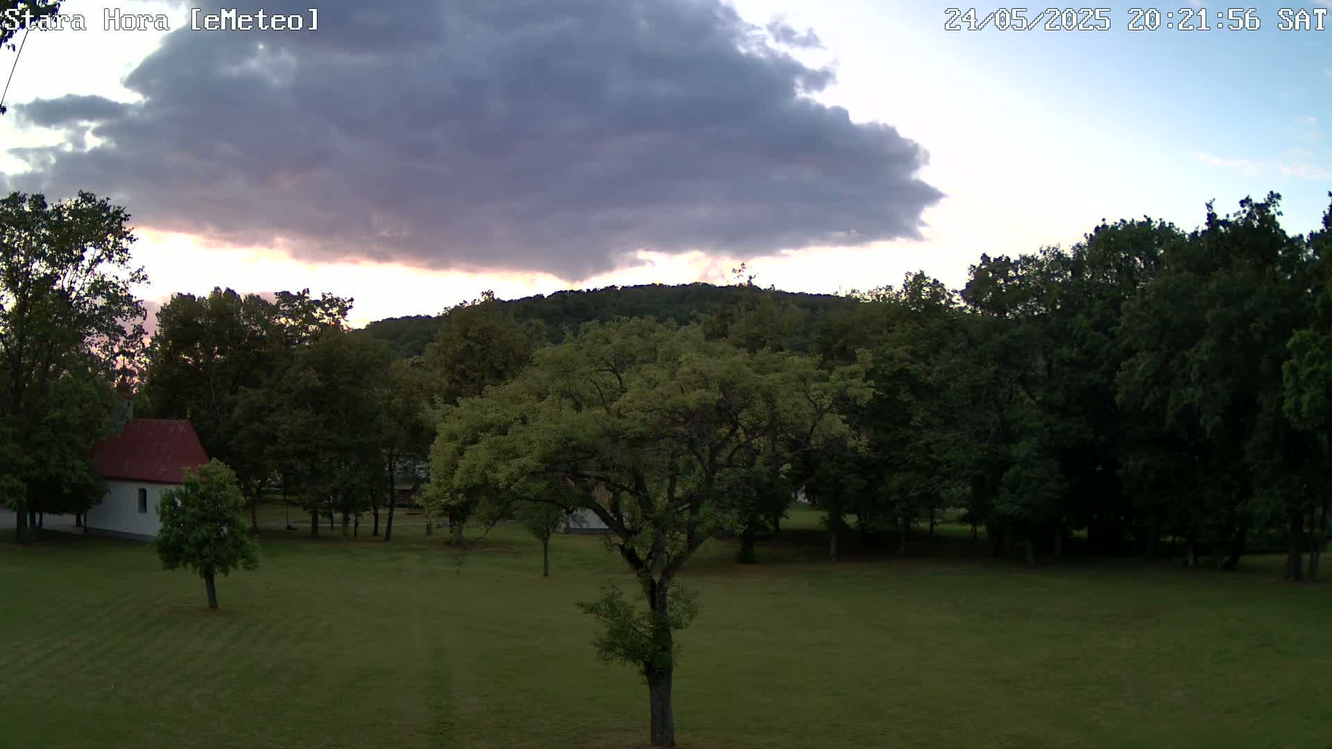 A grassy field with a small building and numerous trees under a partly cloudy twilight sky.