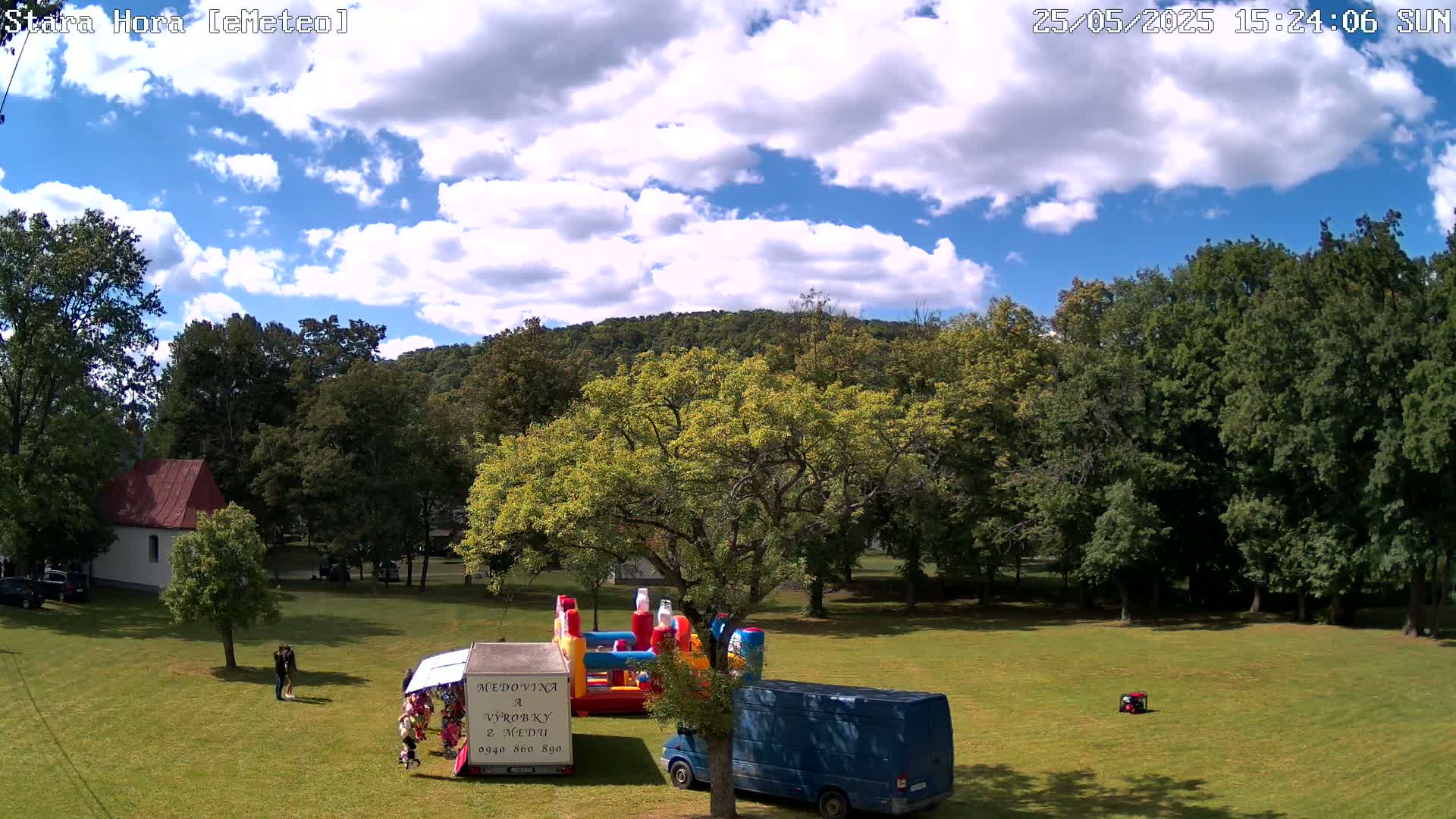 A partly cloudy sunny day shows an open grassy area with inflatable play structures, a blue van, and several people near a small building and trees, with a hill in the background.