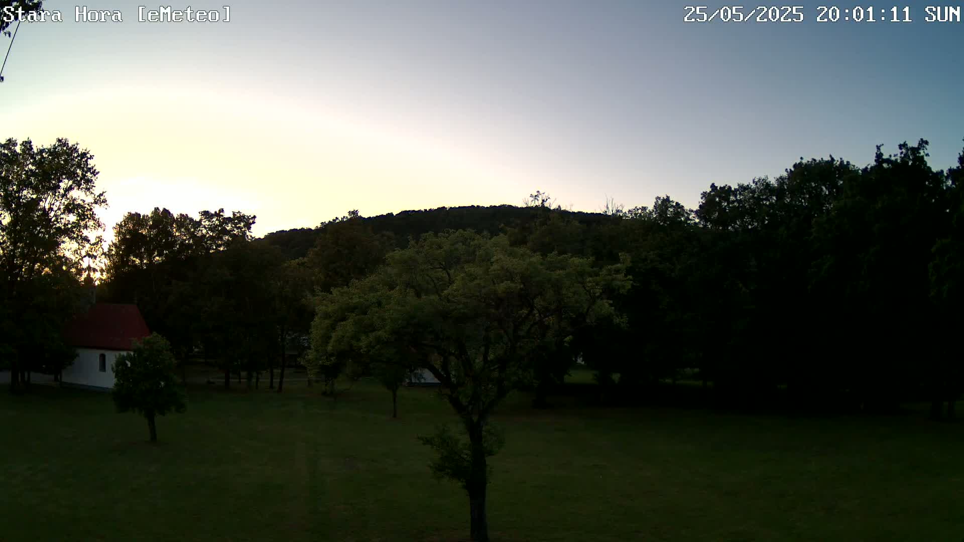 A small white building with a red roof sits on a grassy field surrounded by trees and a hill under a twilight sky.