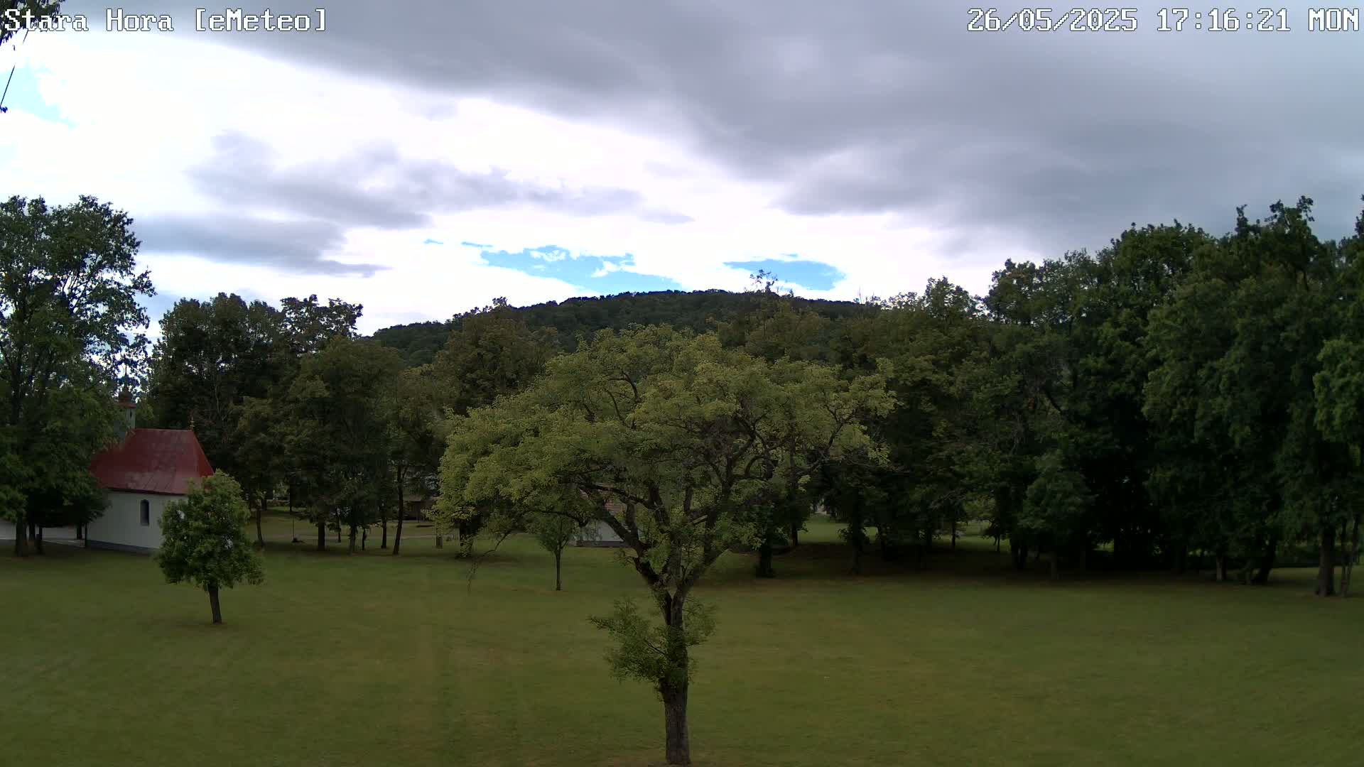 A grassy field with a small building and several trees, under a partly cloudy sky, sits before a line of dark green trees and a hill in the background.