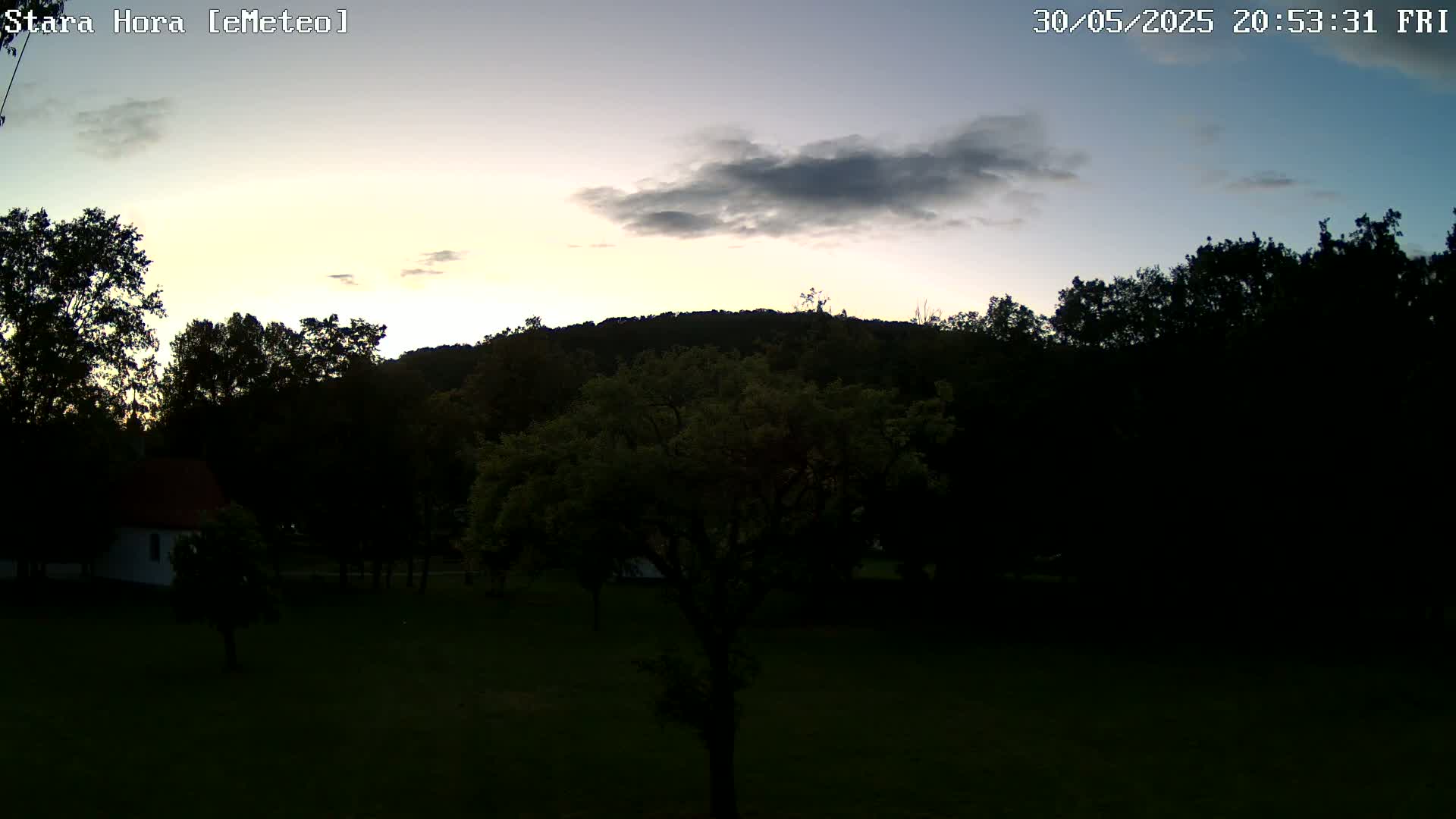 A mostly cloudy twilight sky illuminates a grassy field, trees, and a small house near a hill.