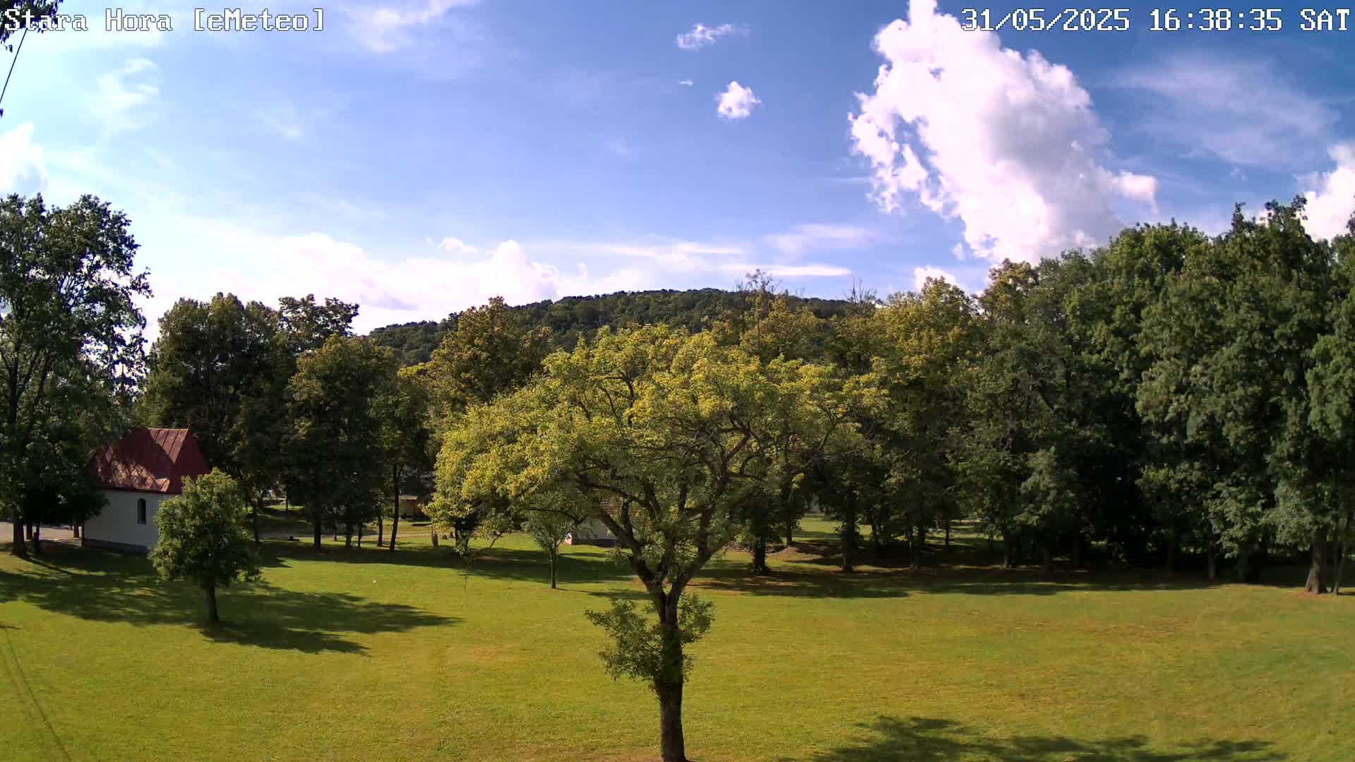 A partly sunny day shows a grassy field with deciduous trees, a small white building with a red roof, and a hill in the background.