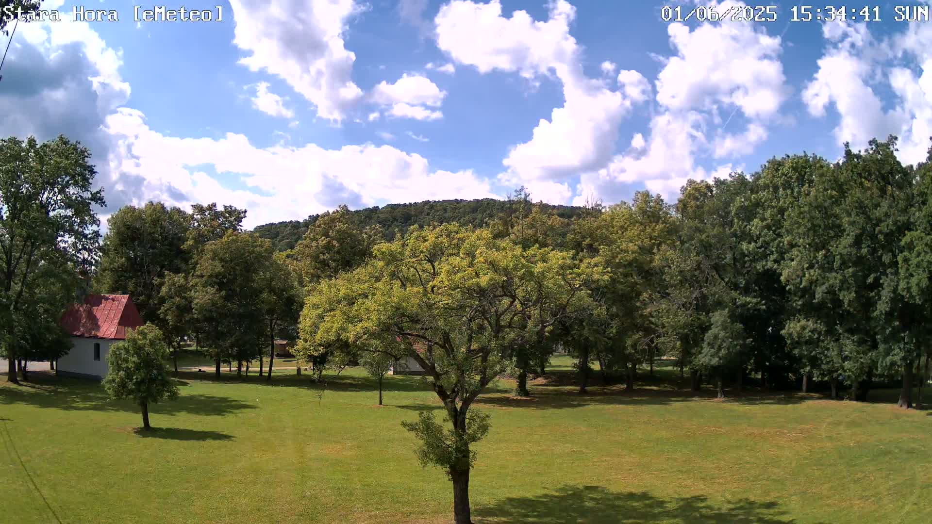 A partly cloudy sunny day reveals a grassy field with a small white building and many green trees, leading up to a hill in the background.