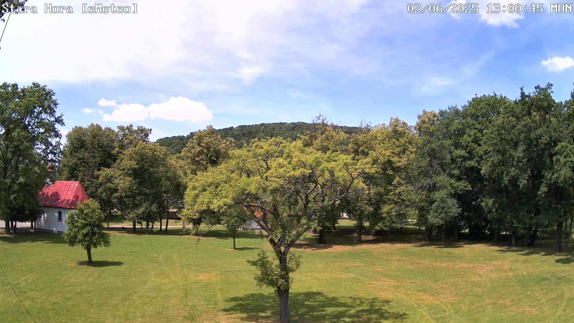 A mostly sunny day reveals a grassy park with deciduous trees surrounding a small, white building with a red roof, backed by a forested hill.