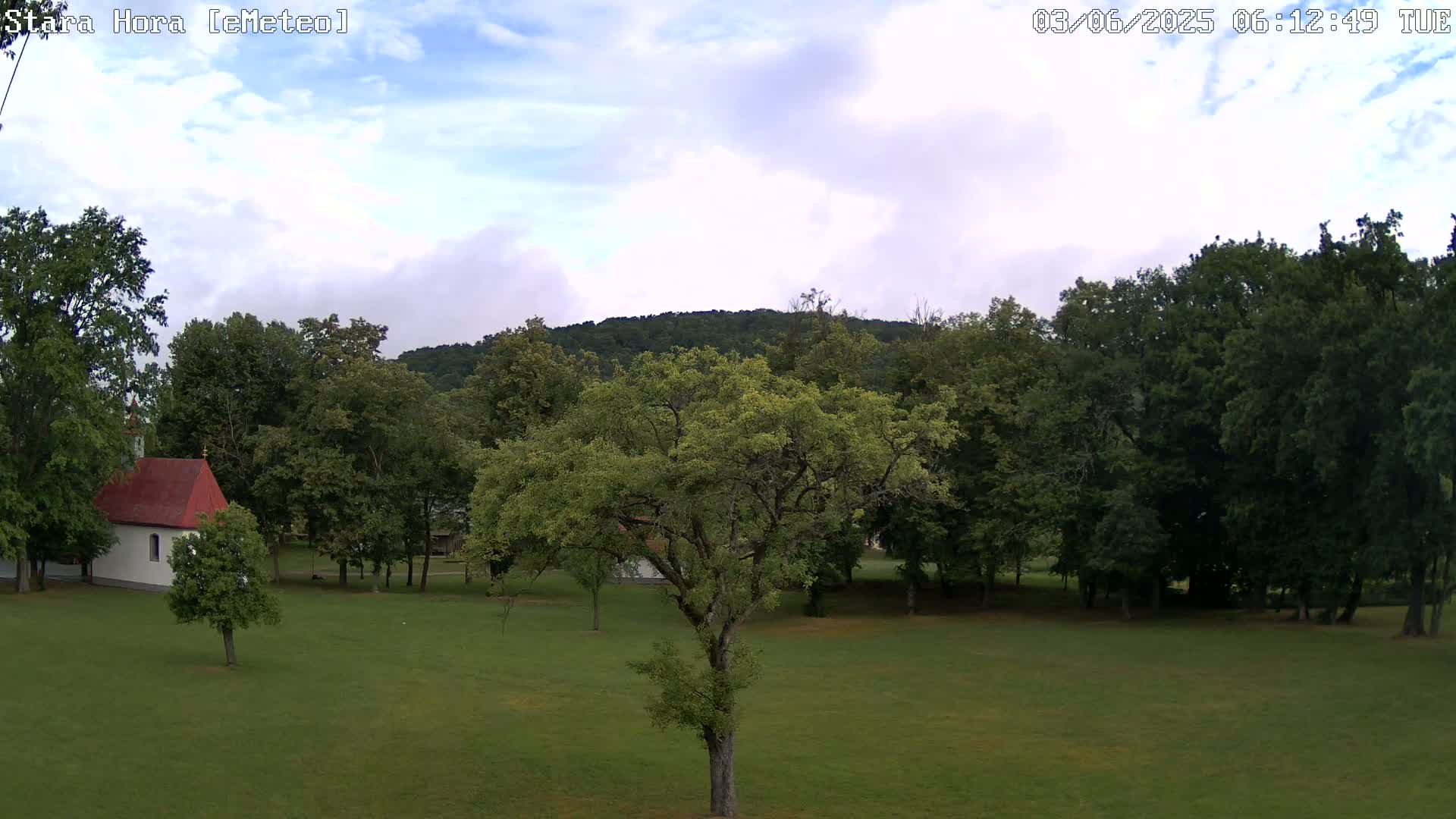 A partly cloudy sky overlooks a grassy field with a small white chapel and several leafy green trees, some of which are large and mature.