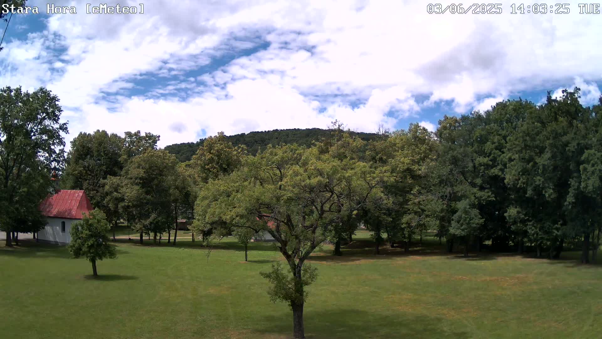 A partly cloudy sky overlooks a grassy field with several trees and a small red-roofed building, surrounded by a larger wooded area and a hill in the background.