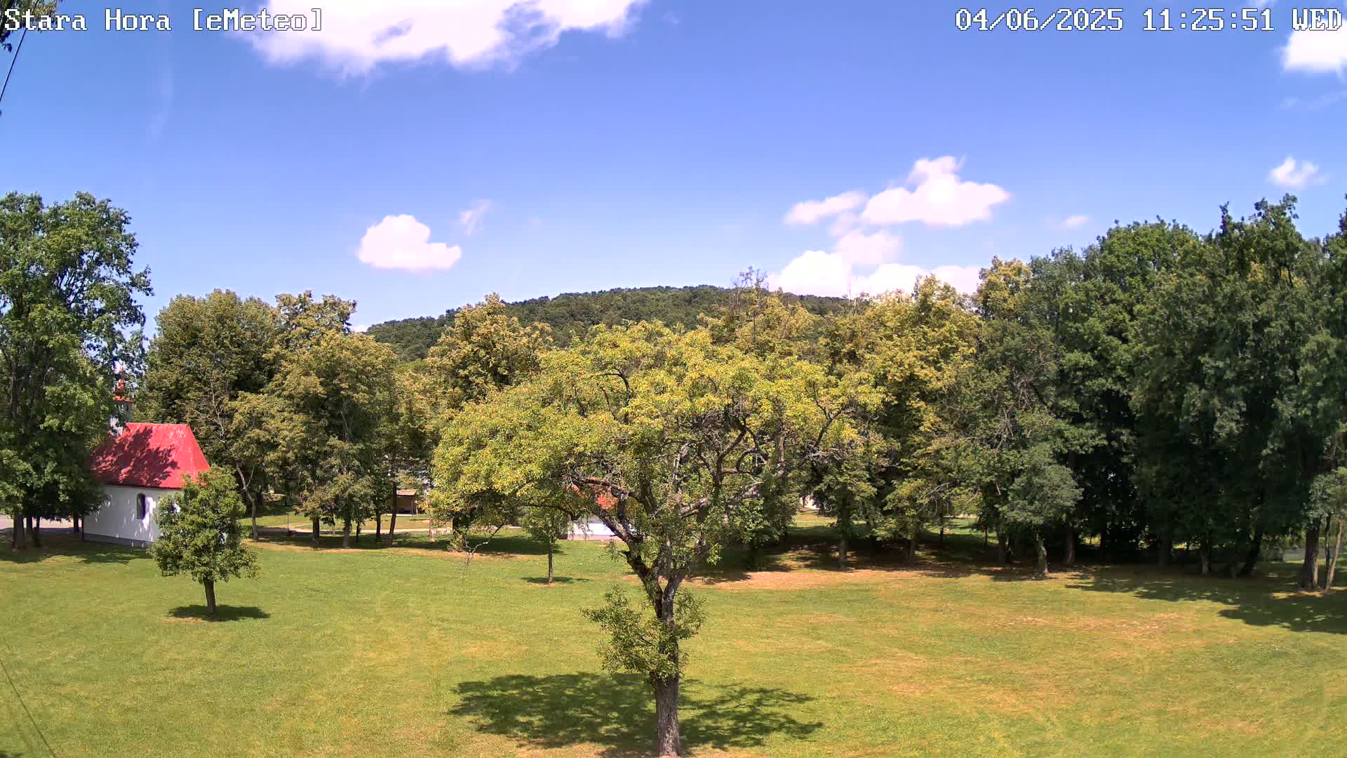 A grassy field with a small white building with a red roof, surrounded by lush green trees under a partly sunny sky.