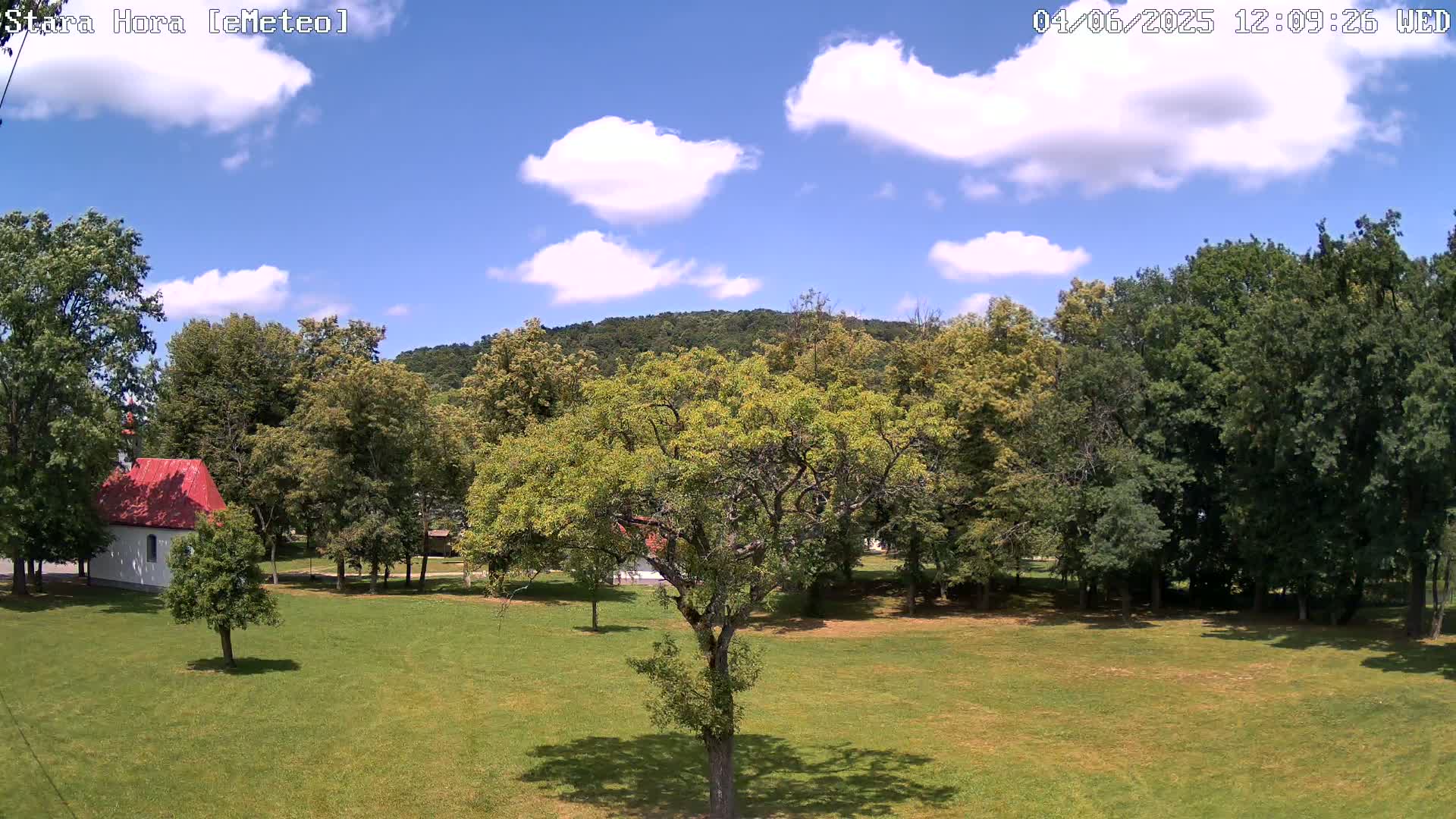 A grassy field with a small red-roofed building and many leafy green trees under a partly cloudy blue sky.