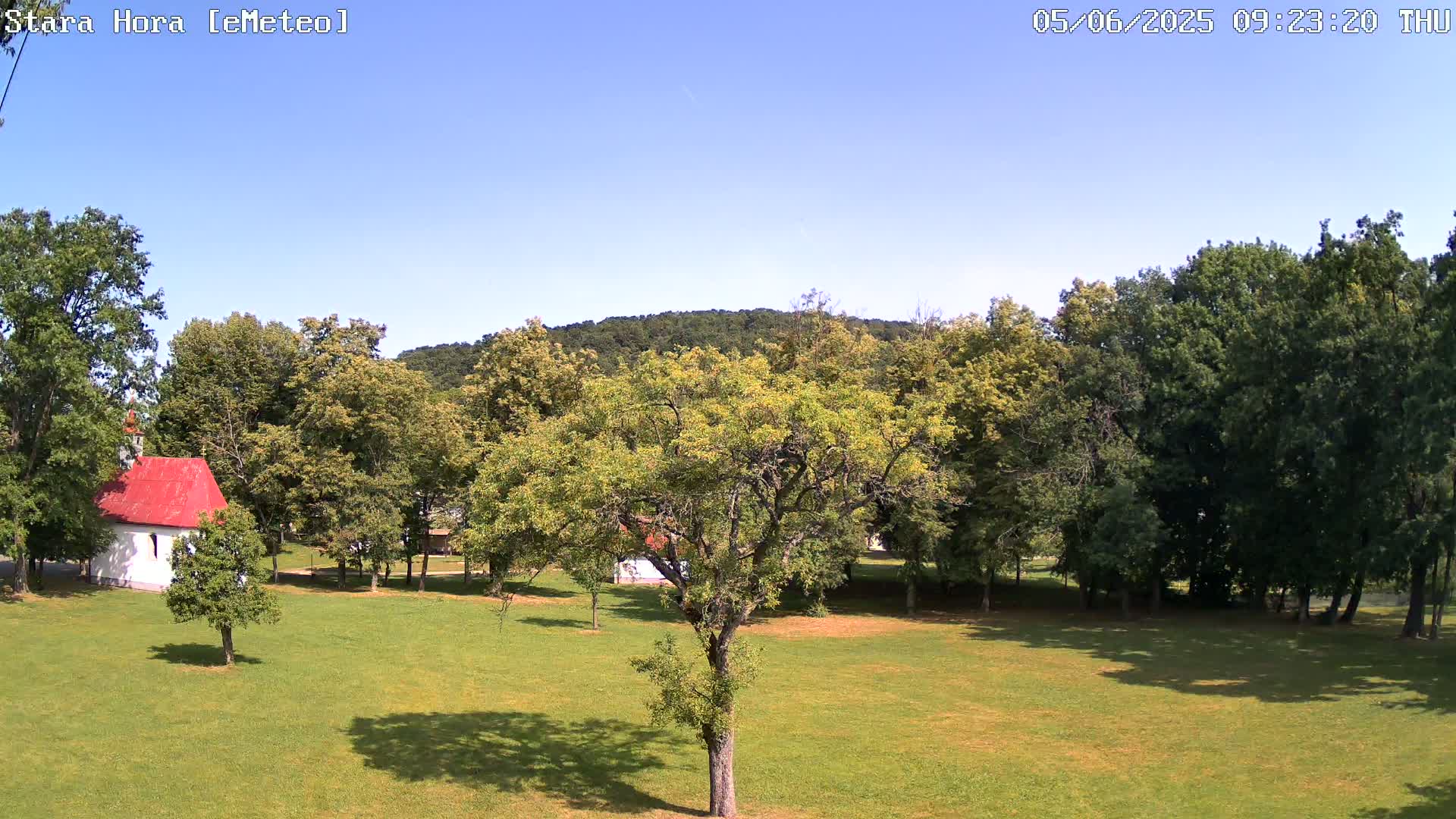 A small white chapel with a red roof sits in a grassy field surrounded by lush green trees under a clear blue sky.