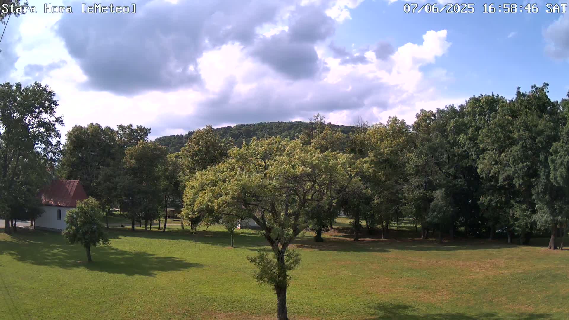 A partly cloudy sky overlooks a grassy field with deciduous trees and a small white building, backed by a tree-covered hill.