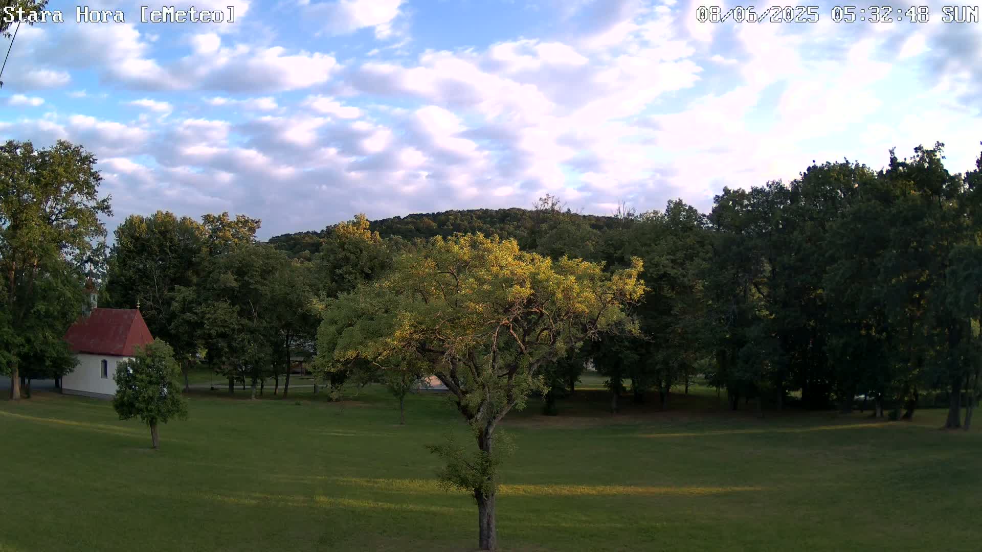 A partly cloudy daytime scene shows a grassy field with a small white building, several trees, and a hill in the background.