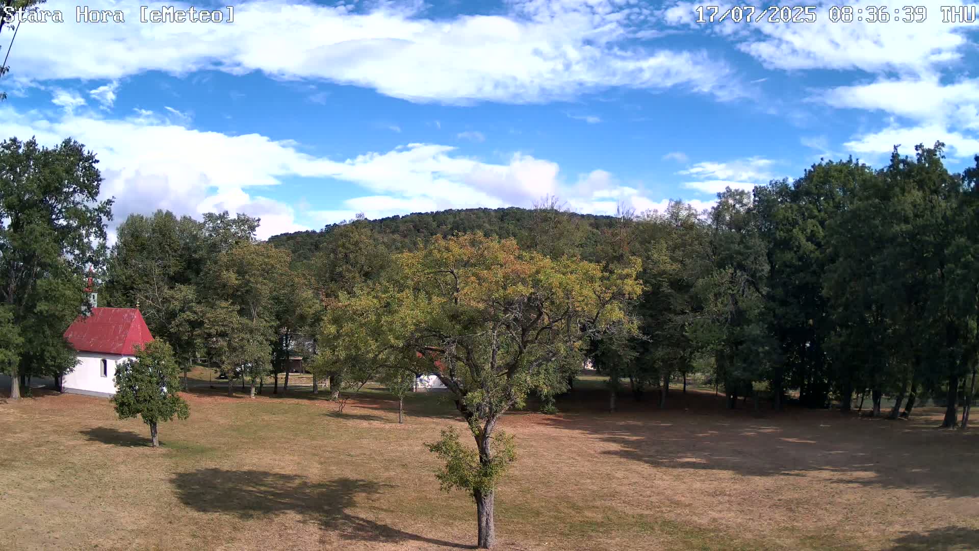 A small white chapel with a red roof sits in a grassy field surrounded by deciduous trees under a partly cloudy blue sky.