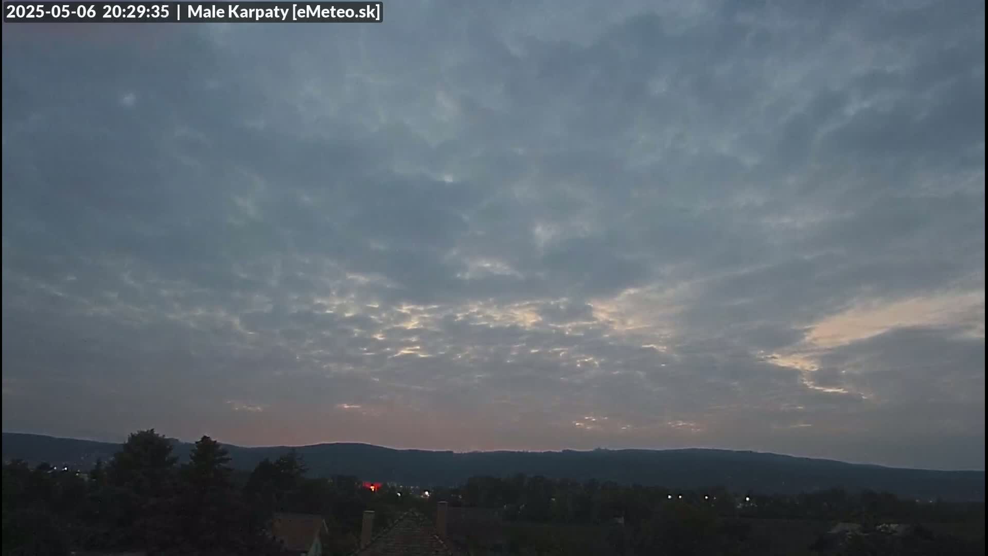 A mostly cloudy twilight sky over a dark, hilly landscape with a few scattered lights visible in the distance.