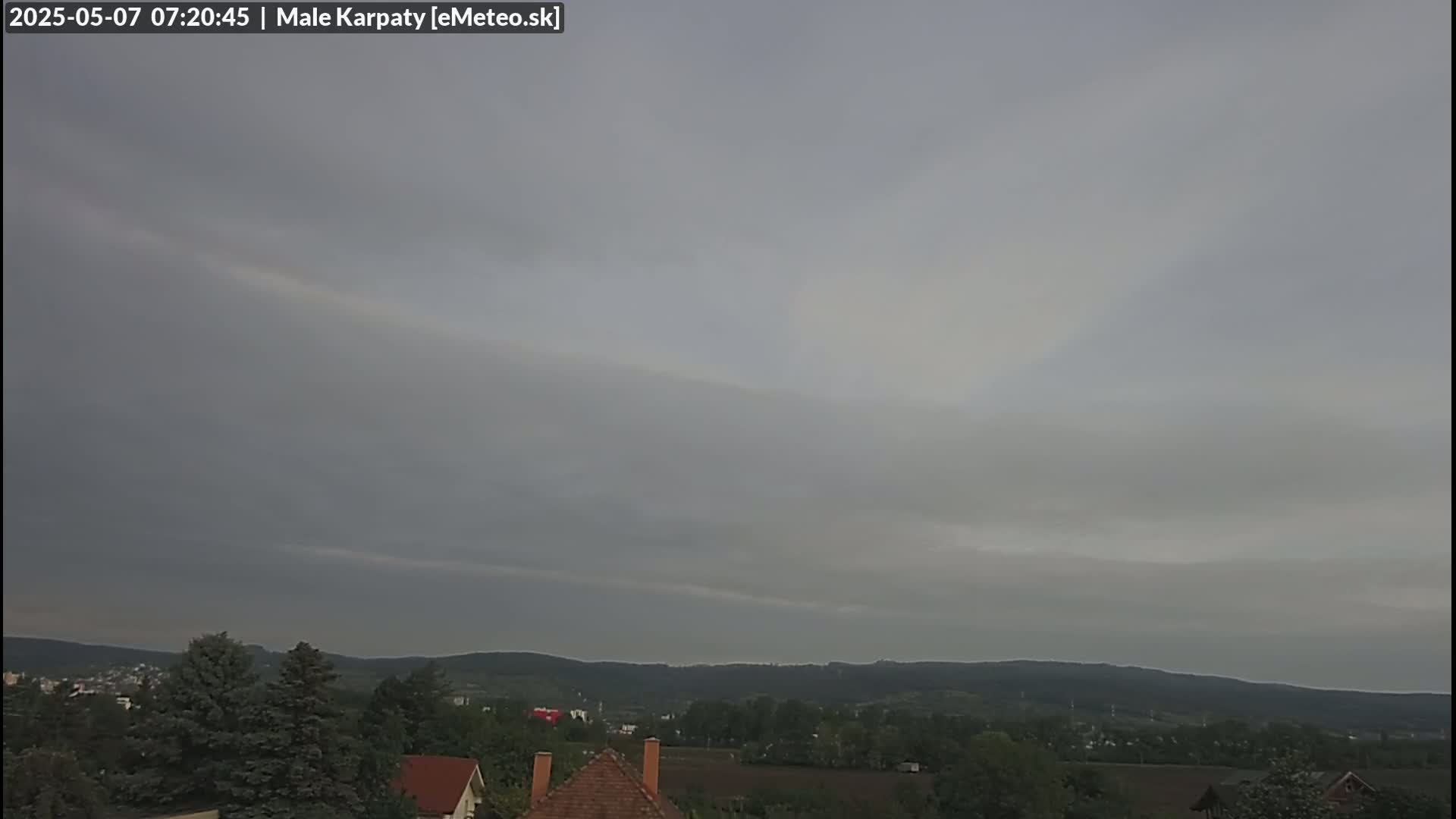 A faint rainbow is visible in an overcast sky above a line of dark green hills and a village with a few houses.